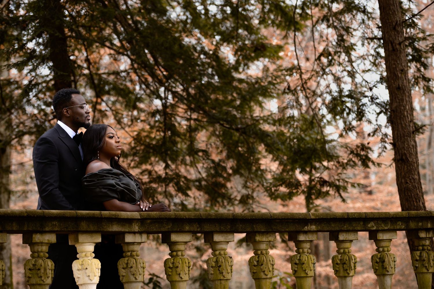 Couple in formal attire sitting on stone balustrade in autumn garden setting.