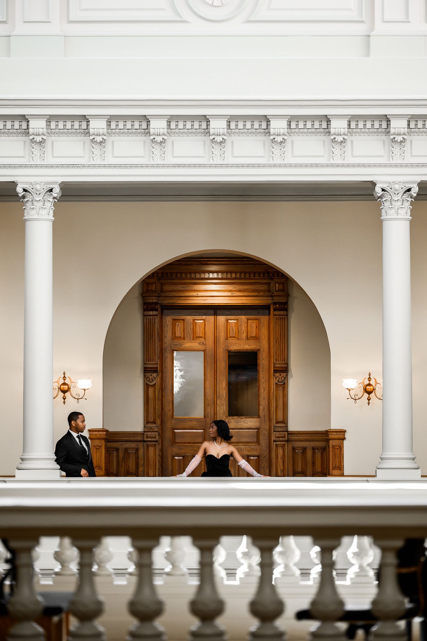 A couple poses separately in an elegant white columned hallway with wooden doors and wall sconces.