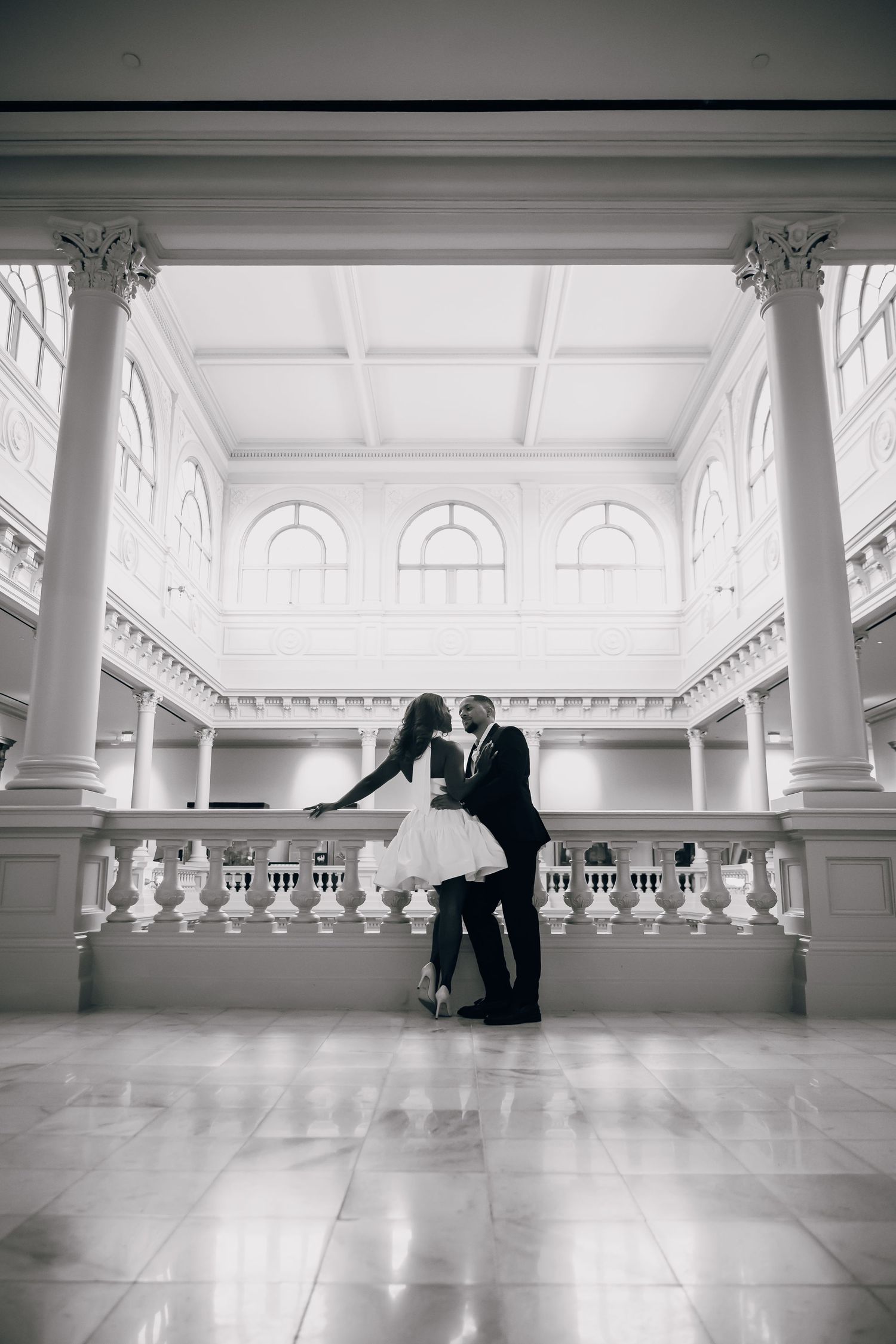 Wide shot of couple dancing in an elegant white hall with classical columns.