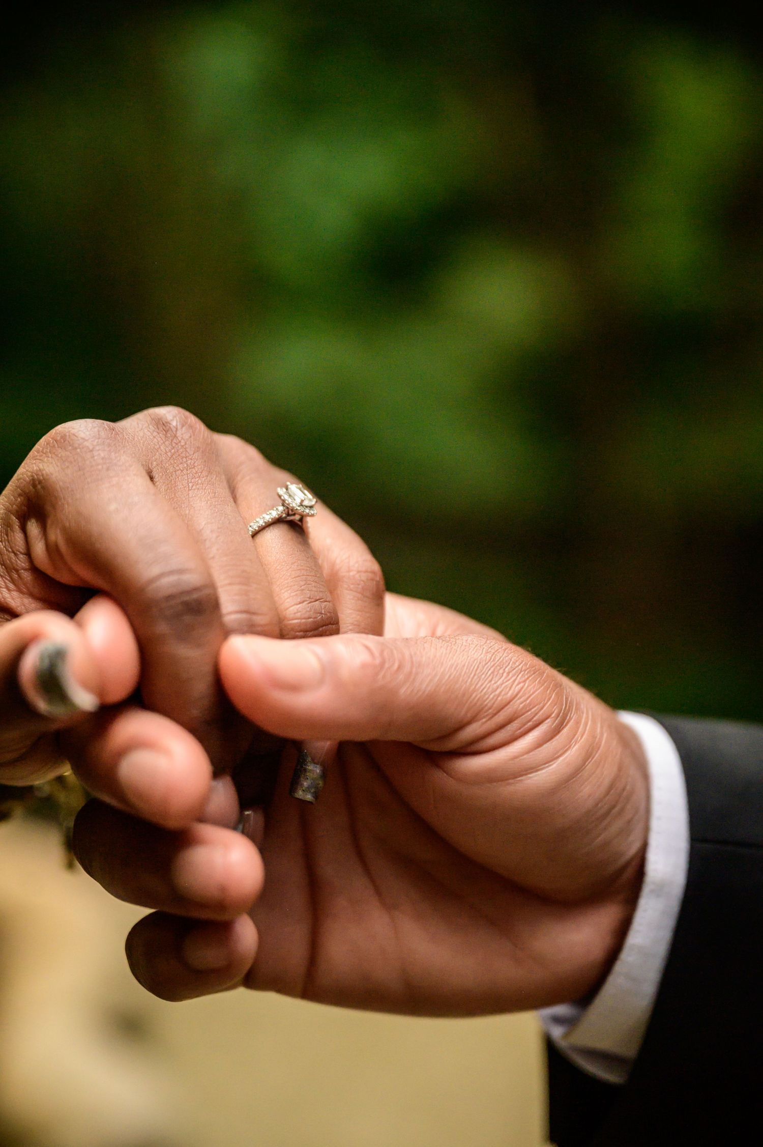 Close-up of hands displaying elegant engagement ring against dark formal attire.
