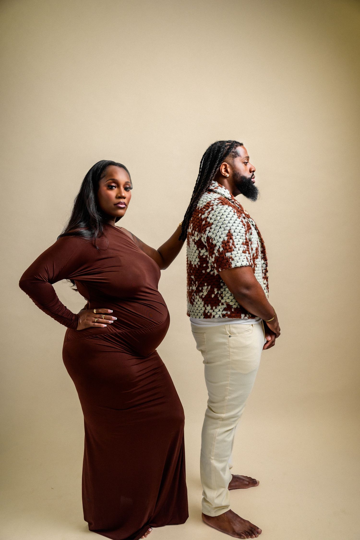 Artistic maternity session with couple in brown dress and white pants against beige backdrop.