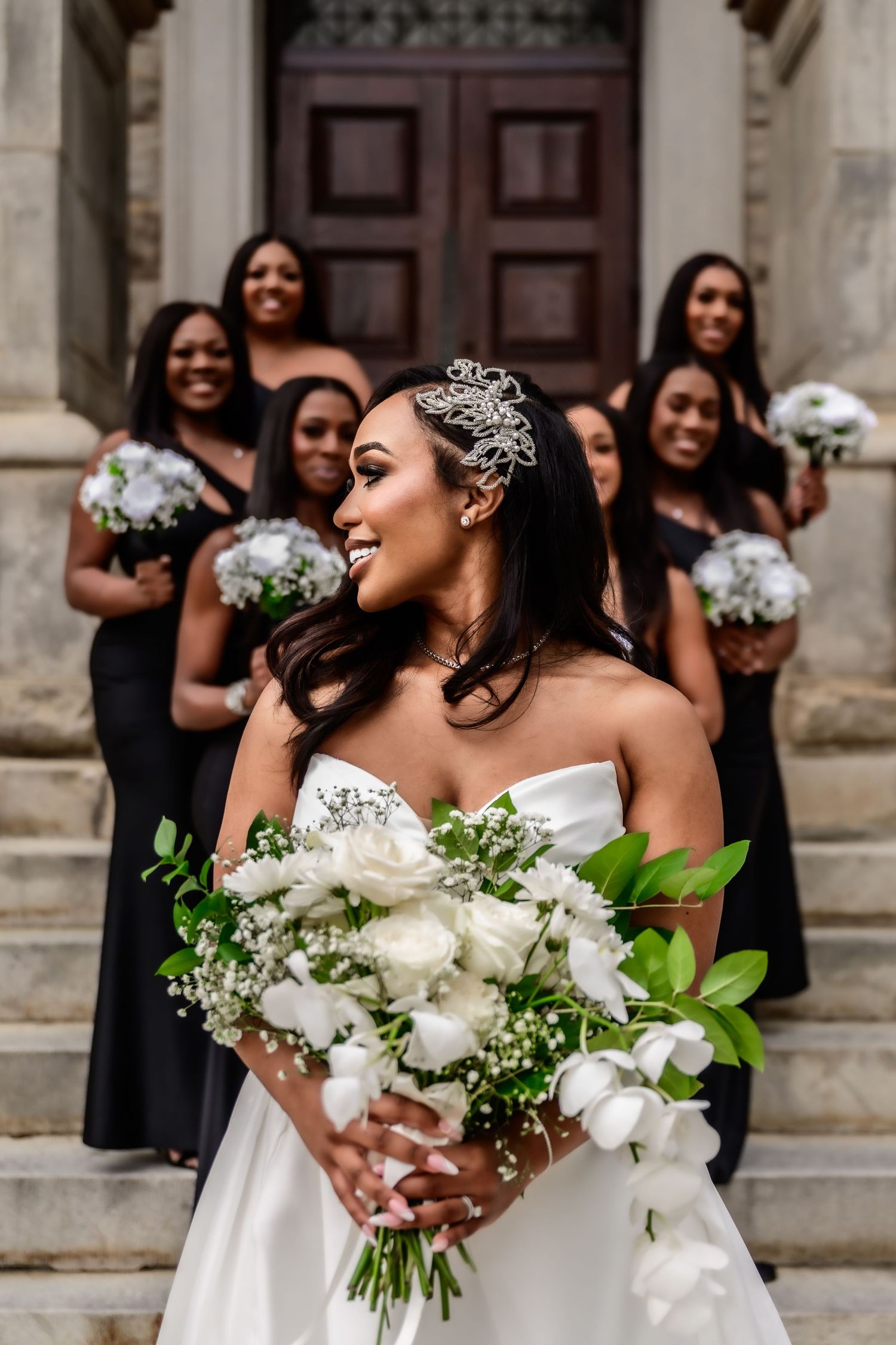 Bridesmaids in black dresses pose with white floral bouquets outside a historic stone building.