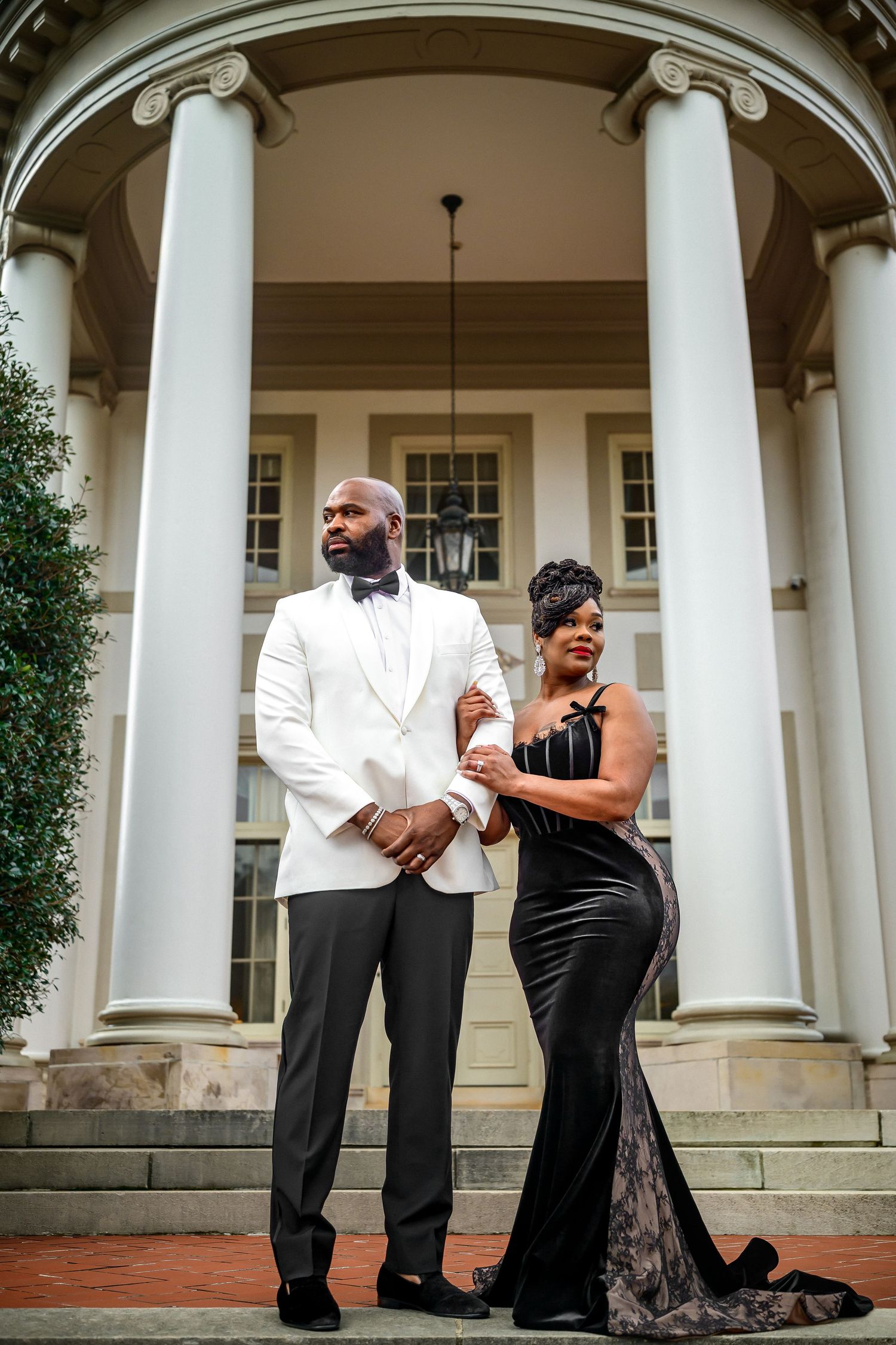 Elegant couple in formal attire pose together in front of grand white columns at a historic building entrance.