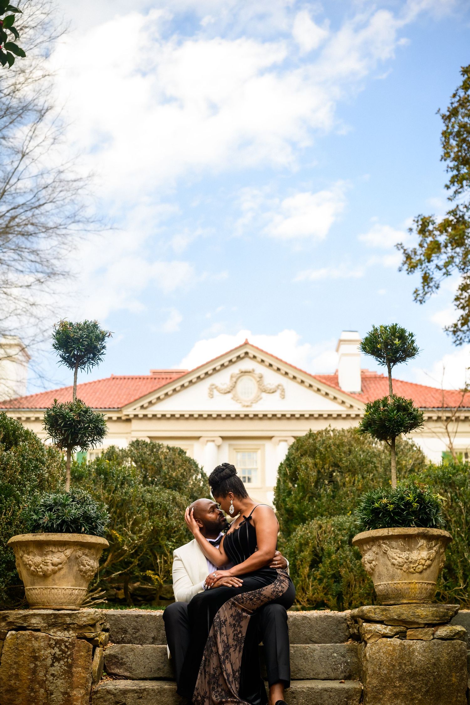Romantic couple embraces on stone wall with Mediterranean villa and garden setting under blue sky.