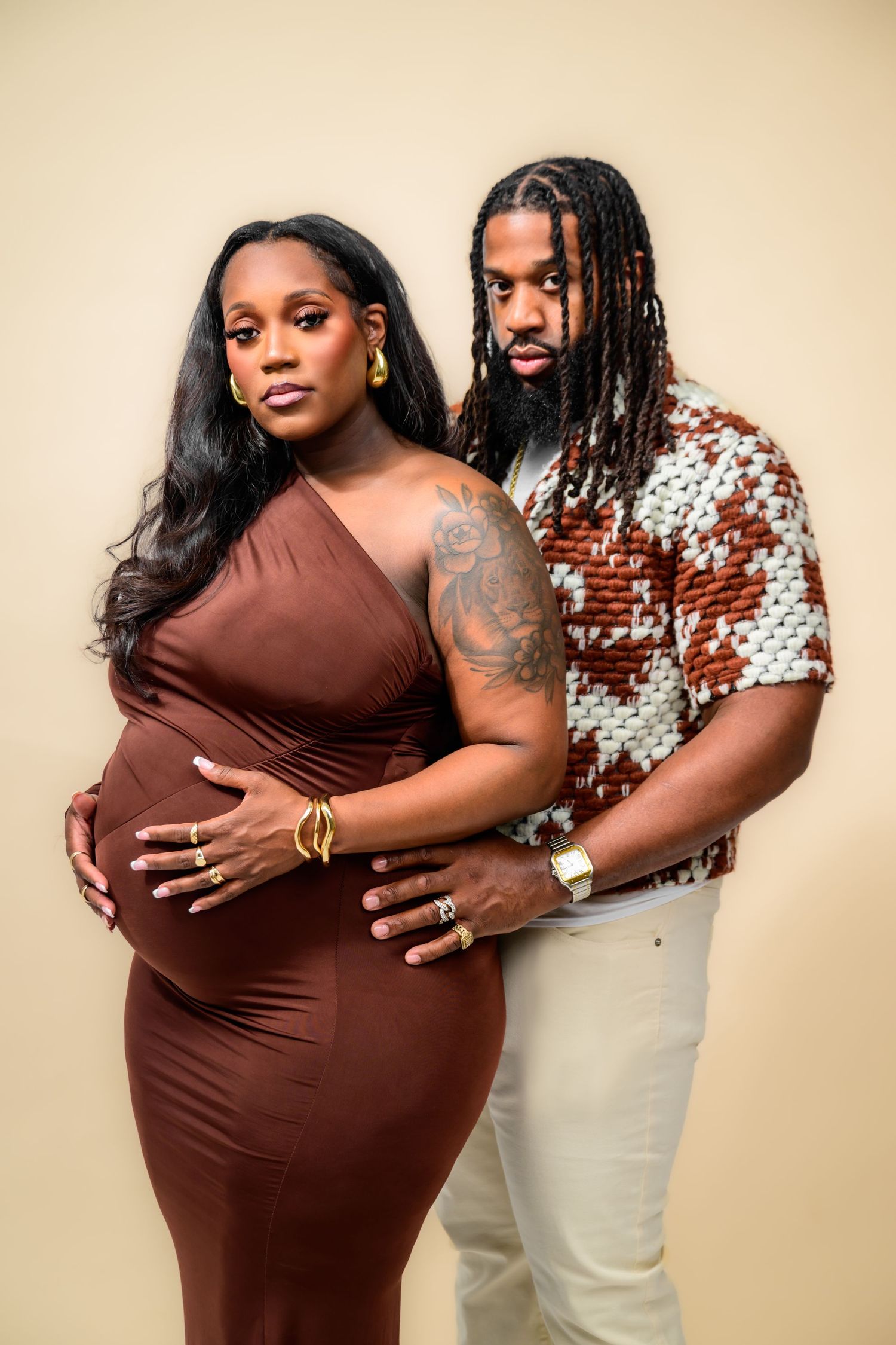 A maternity photoshoot featuring a couple embracing against a beige backdrop.