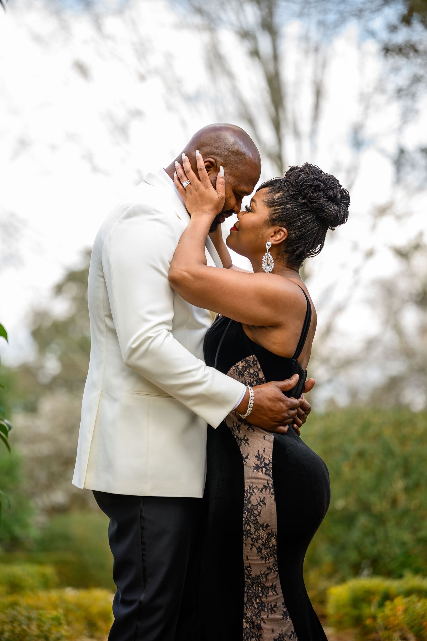 A romantic couple shares a tender embrace and kiss in an outdoor garden setting with blurred foliage in the background.