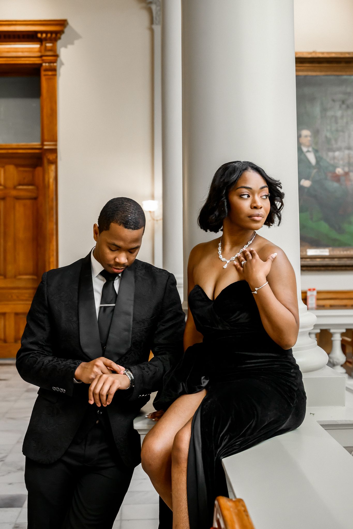 Formal couple in black attire posing intimately against classical indoor architecture.