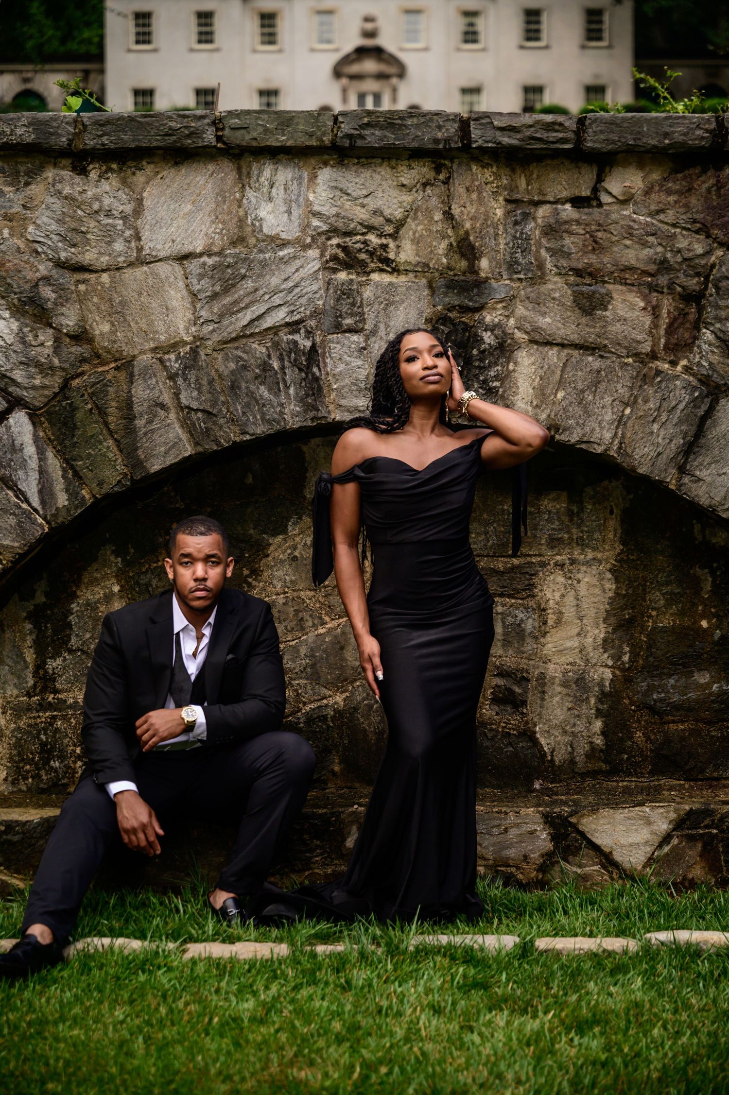 Couple in formal attire pose dramatically against stone wall and grass backdrop.