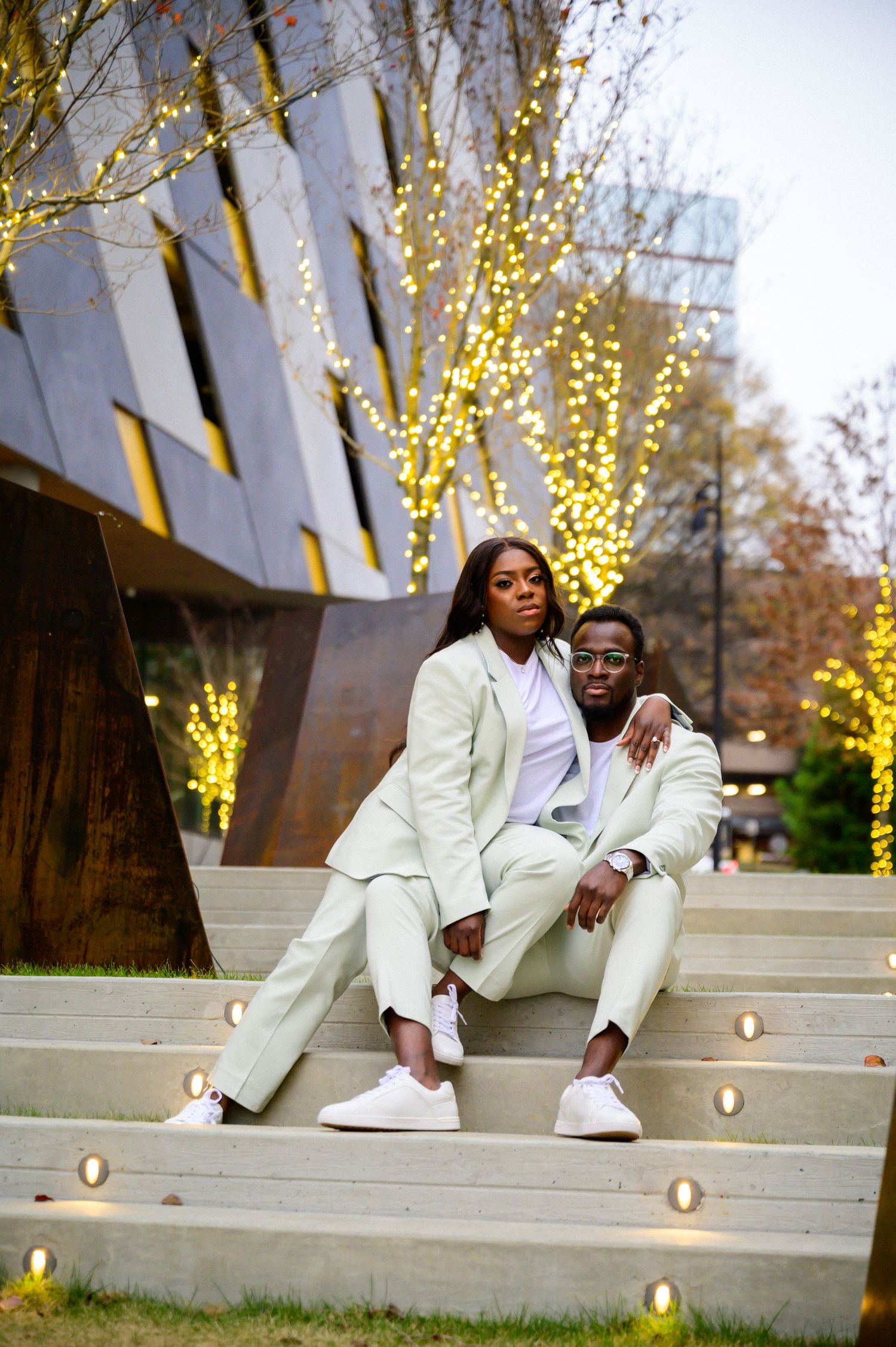Couple relaxes on illuminated concrete steps in matching white formal attire at dusk.