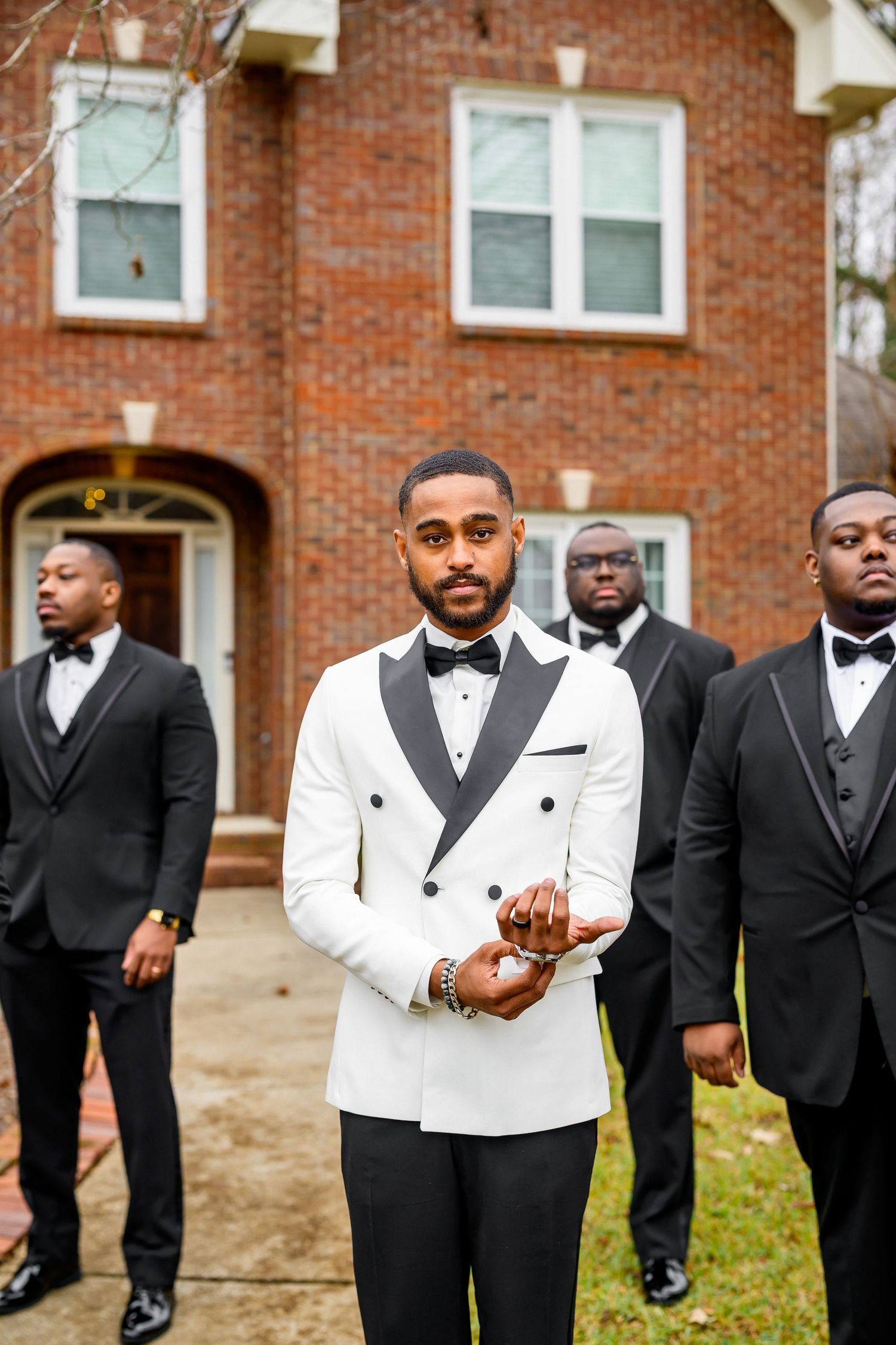 Groomsmen in black and white tuxedos stand together outside a brick building during a formal wedding event.