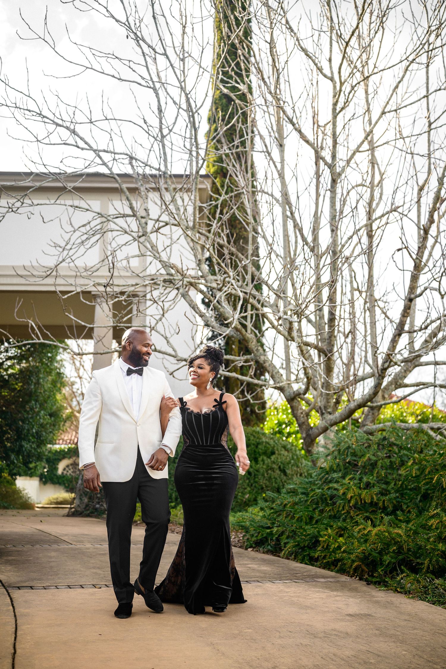 A couple in formal attire shares a romantic moment while walking together on a garden path.