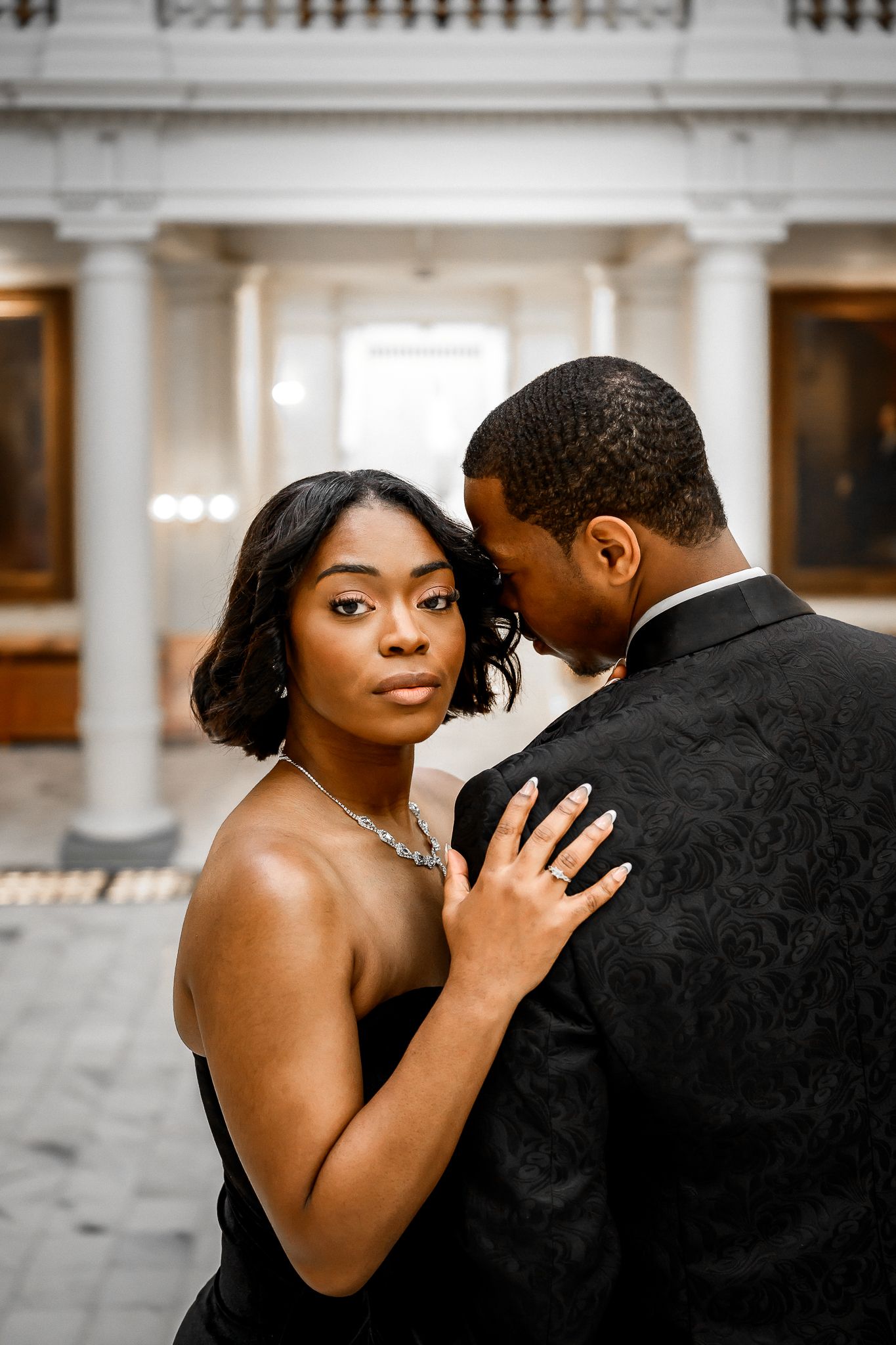 Couple in formal attire embracing in an elegant indoor setting with white columns.