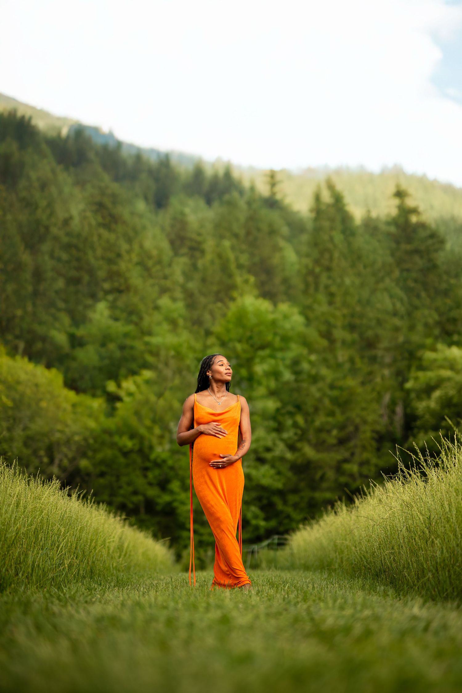 A person in a flowing orange dress stands joyfully in a green meadow with evergreen trees in the background.