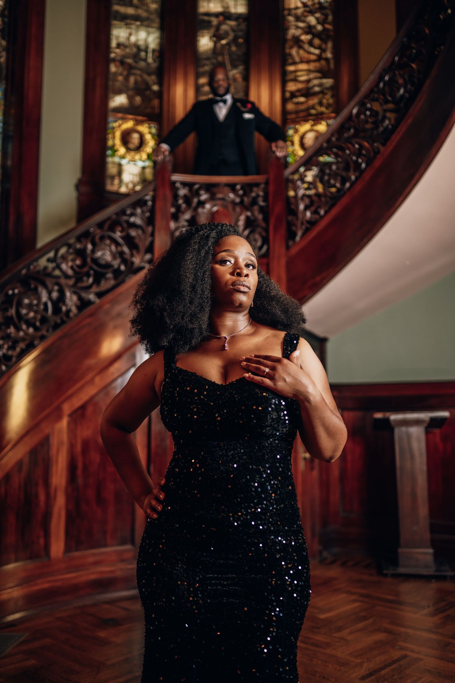 Person in sparkling black gown poses dramatically on ornate wooden staircase.
