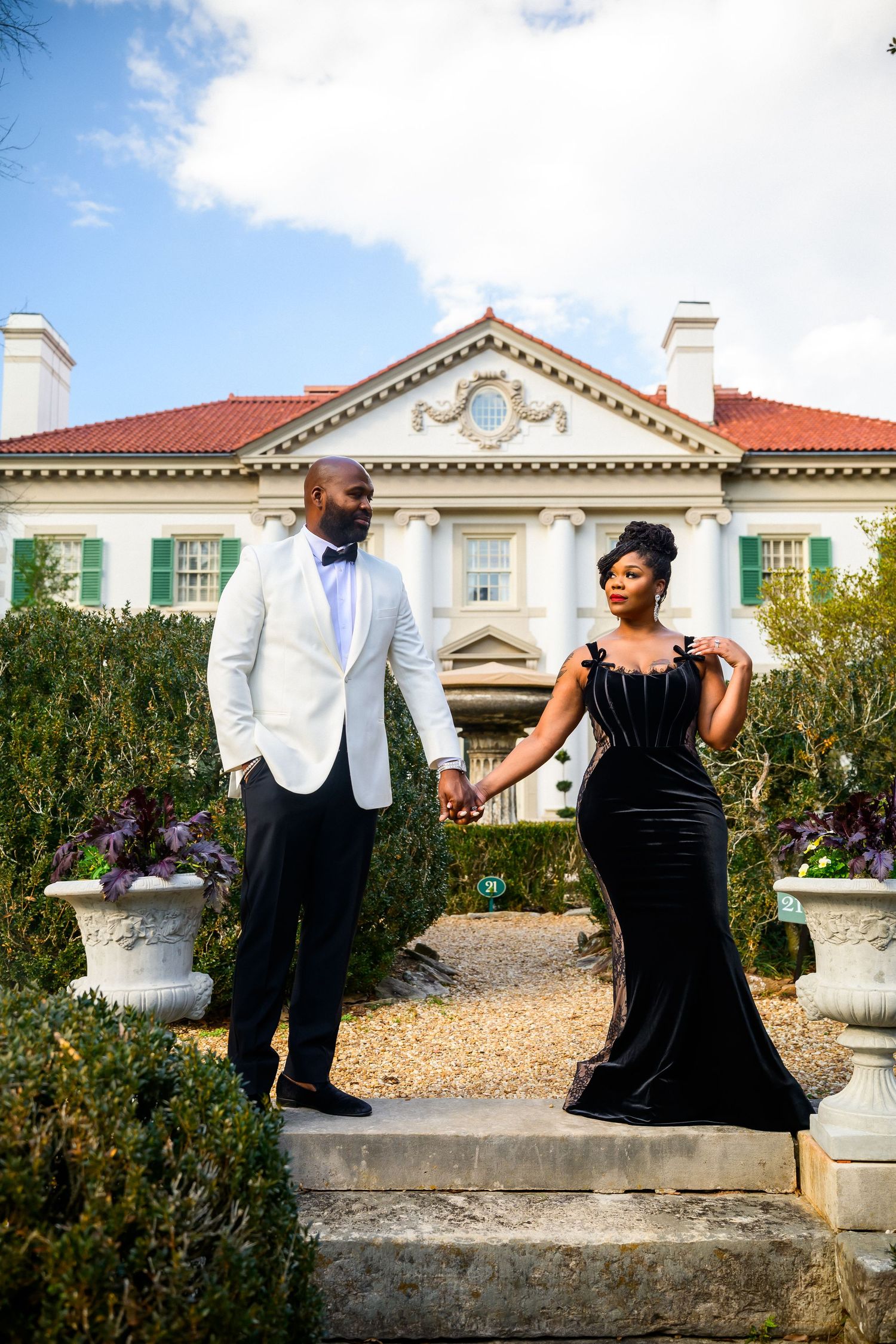 Elegant couple in formal attire pose on stone steps in front of a grand mansion with white columns and red roof.