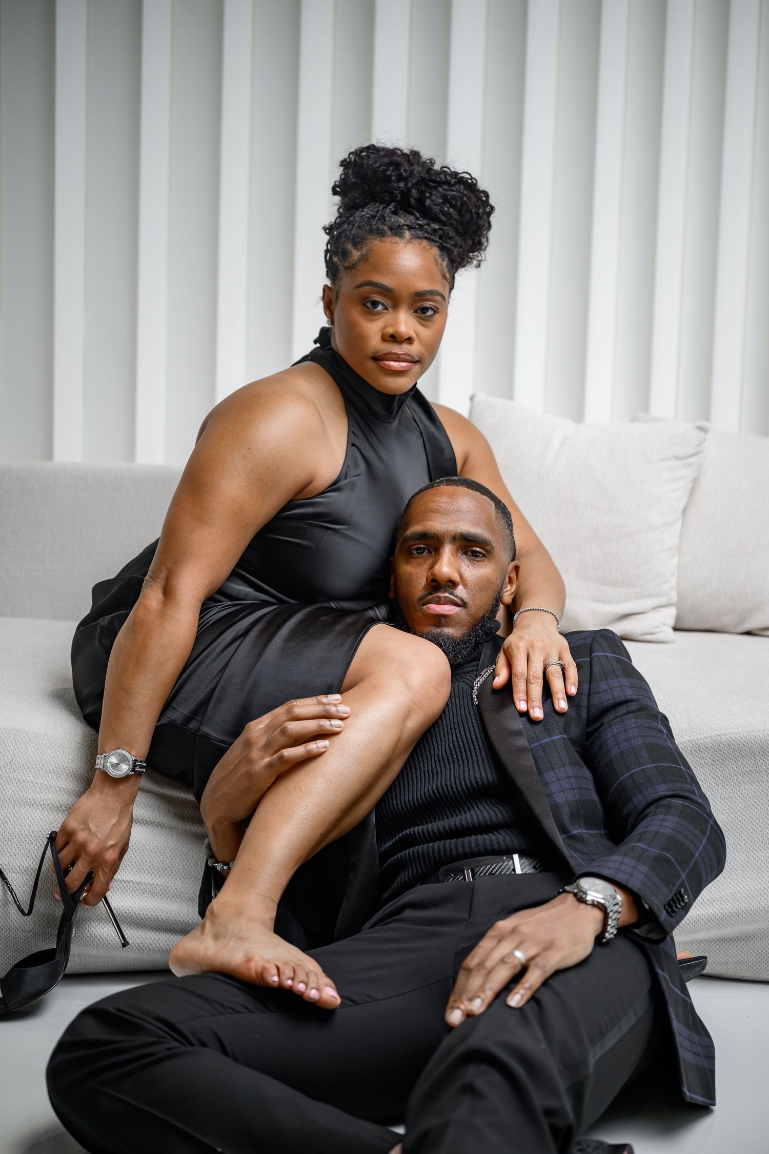 A couple in black outfits poses intimately across a white couch in a modern studio setting.