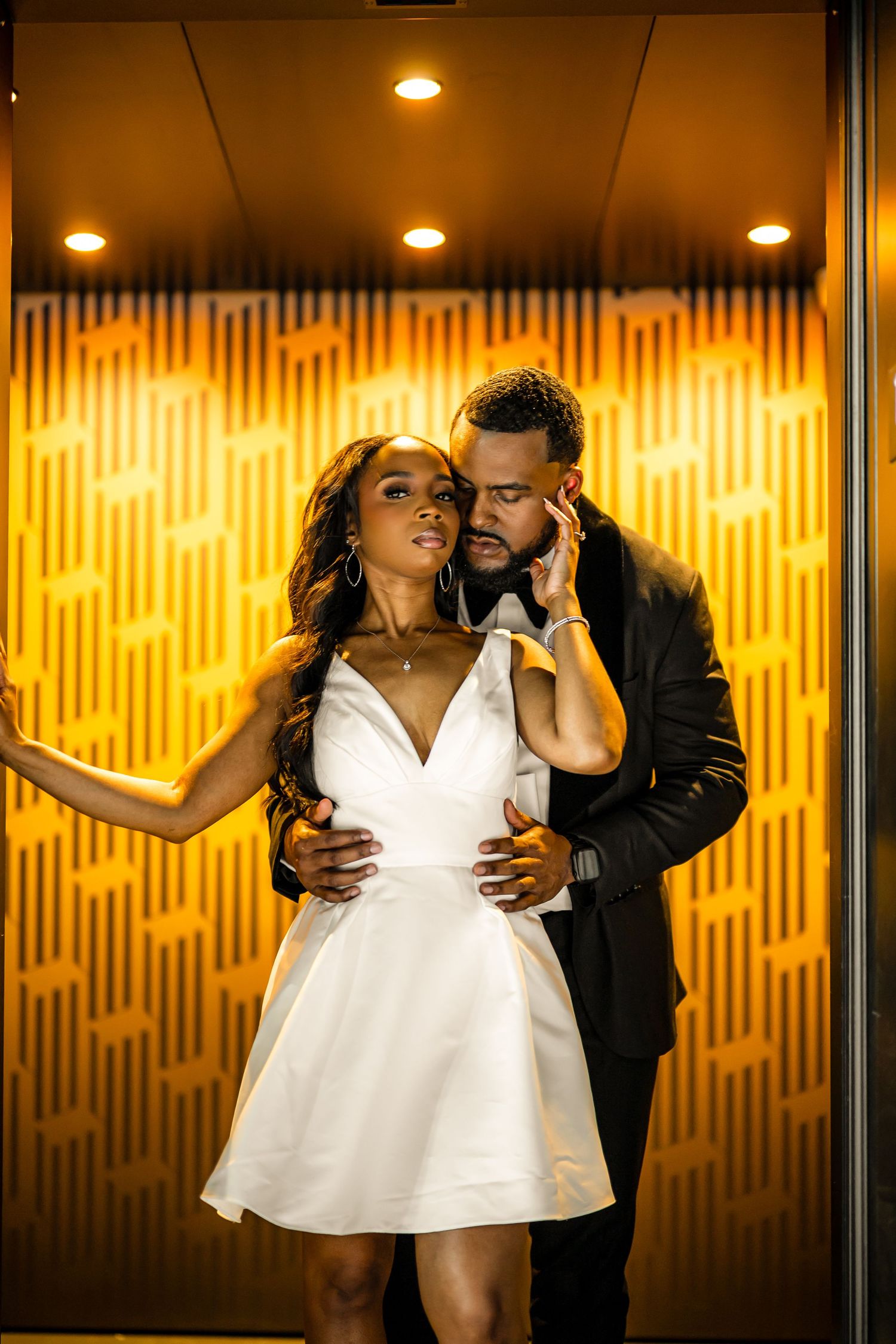 A romantic couple embraces in front of a warm glowing orange wall with recessed lighting.