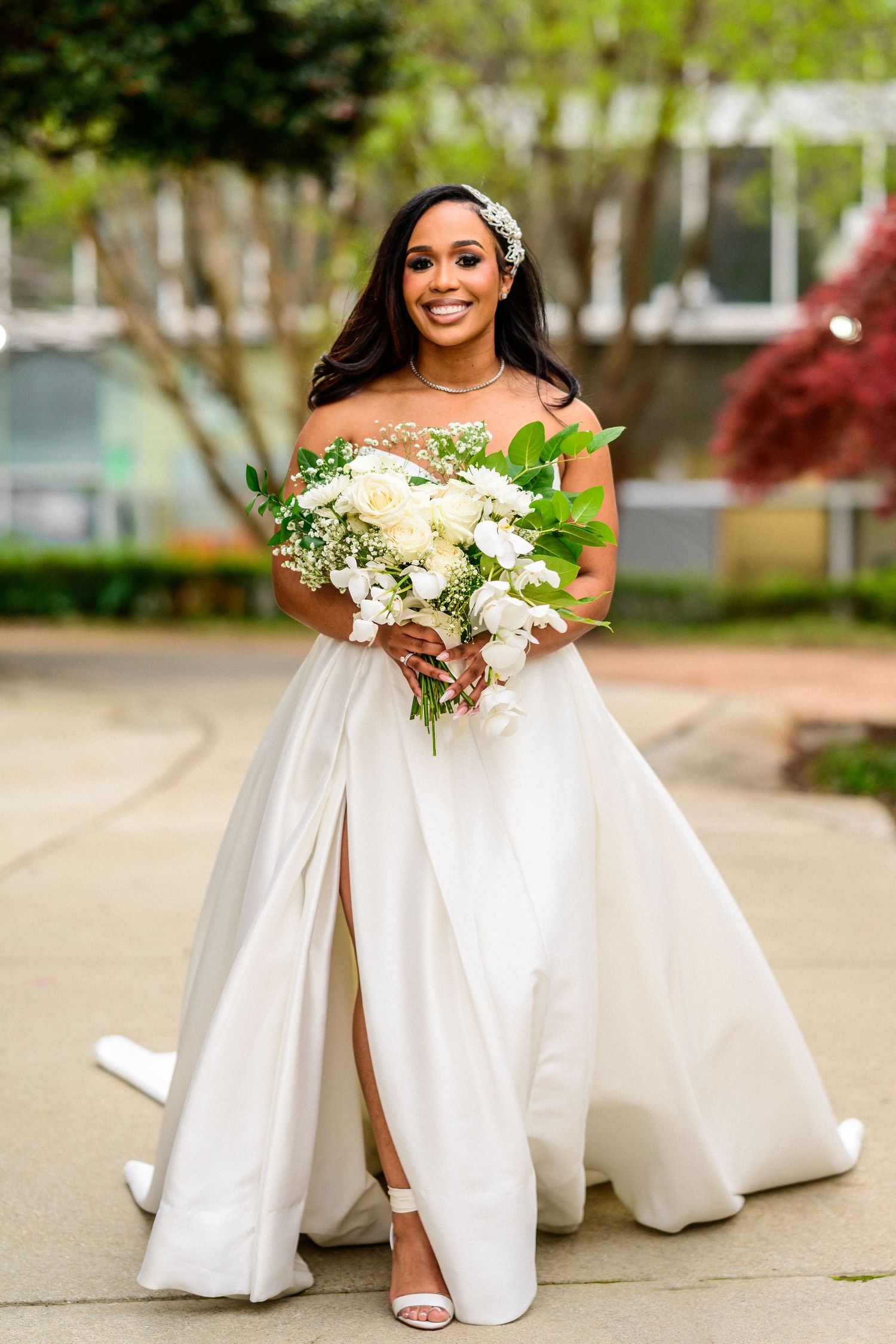 Bride in flowing white gown holds bouquet while sitting on outdoor steps.