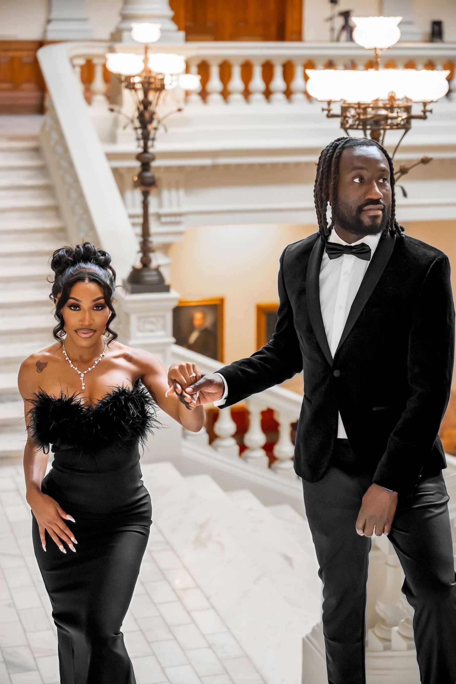Couple in black formal wear posing playfully on elegant marble staircase.