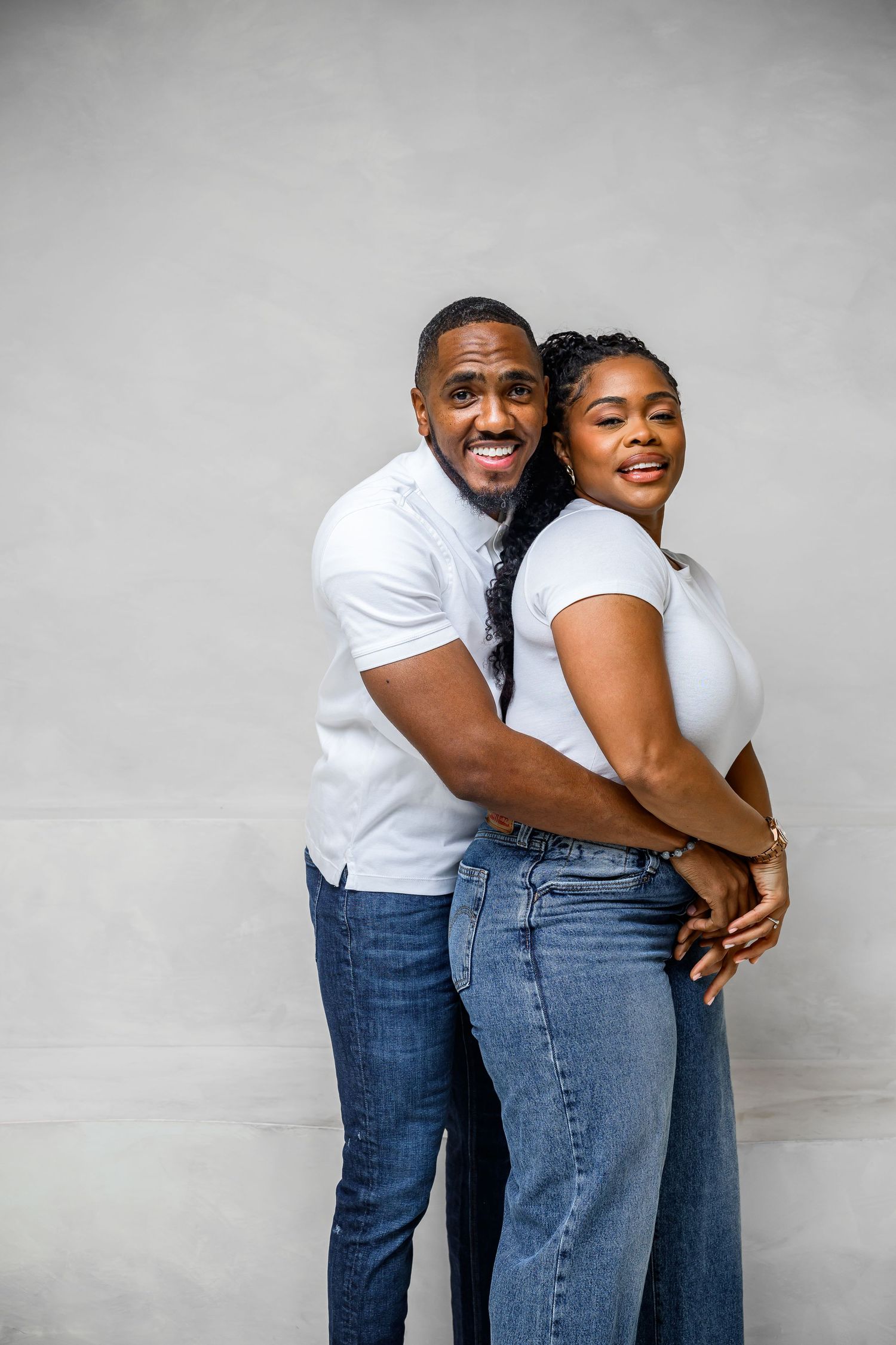 A couple wearing matching white shirts and blue jeans embrace lovingly against a gray background in a series of photos.
