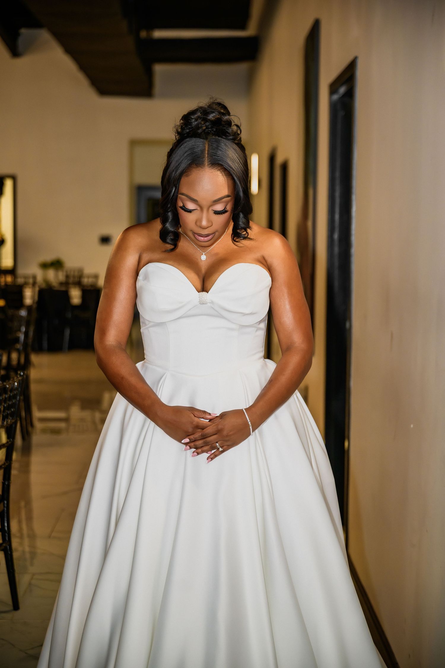 A bride in a stunning white strapless wedding gown poses in an elegant indoor hallway setting.
