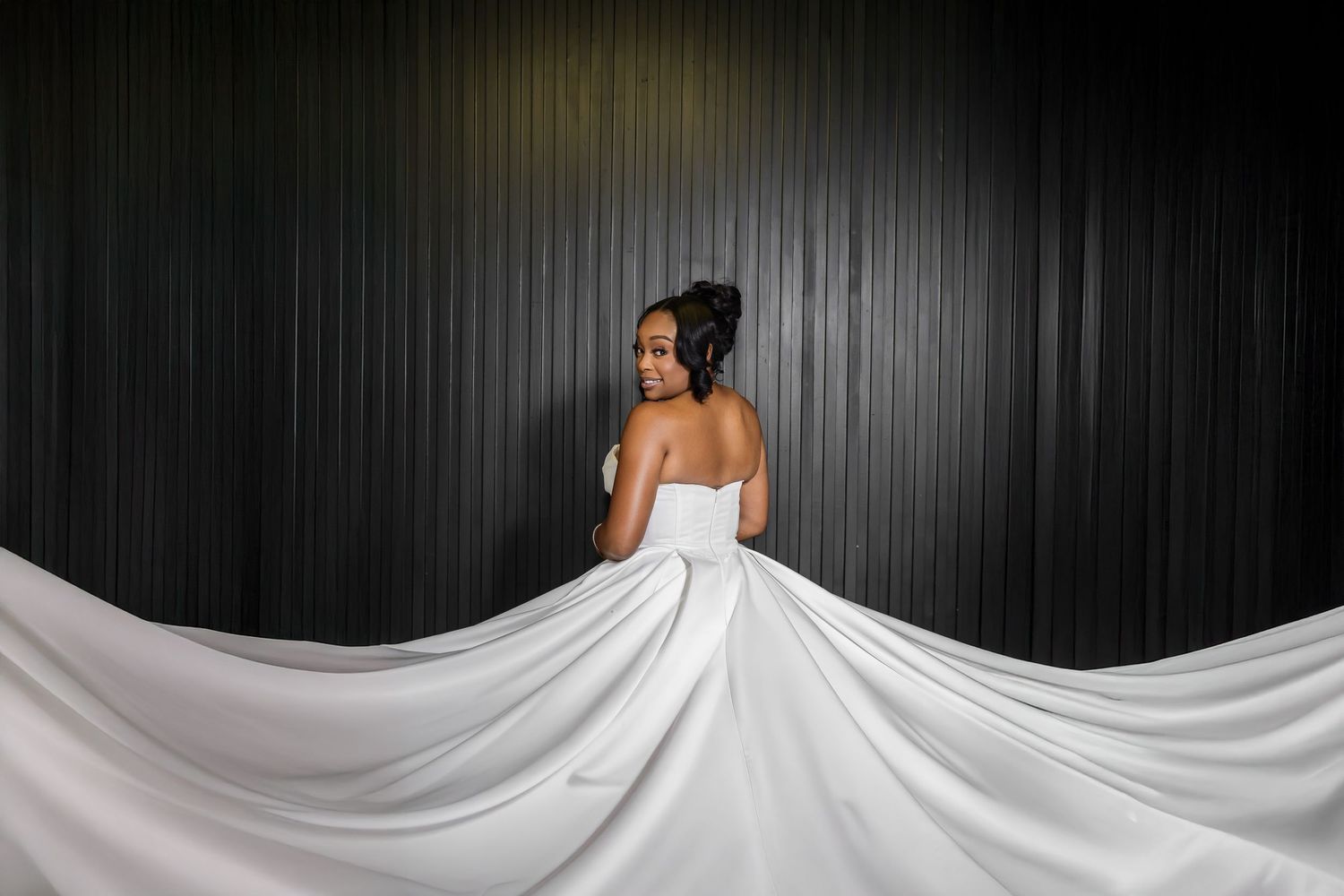 A white wedding dress train flows elegantly across a dark floor in dramatic lighting.