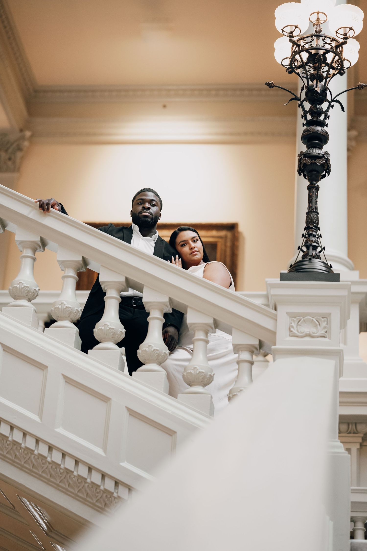 Wedding couple posing on ornate white marble staircase with decorative iron light fixtures.