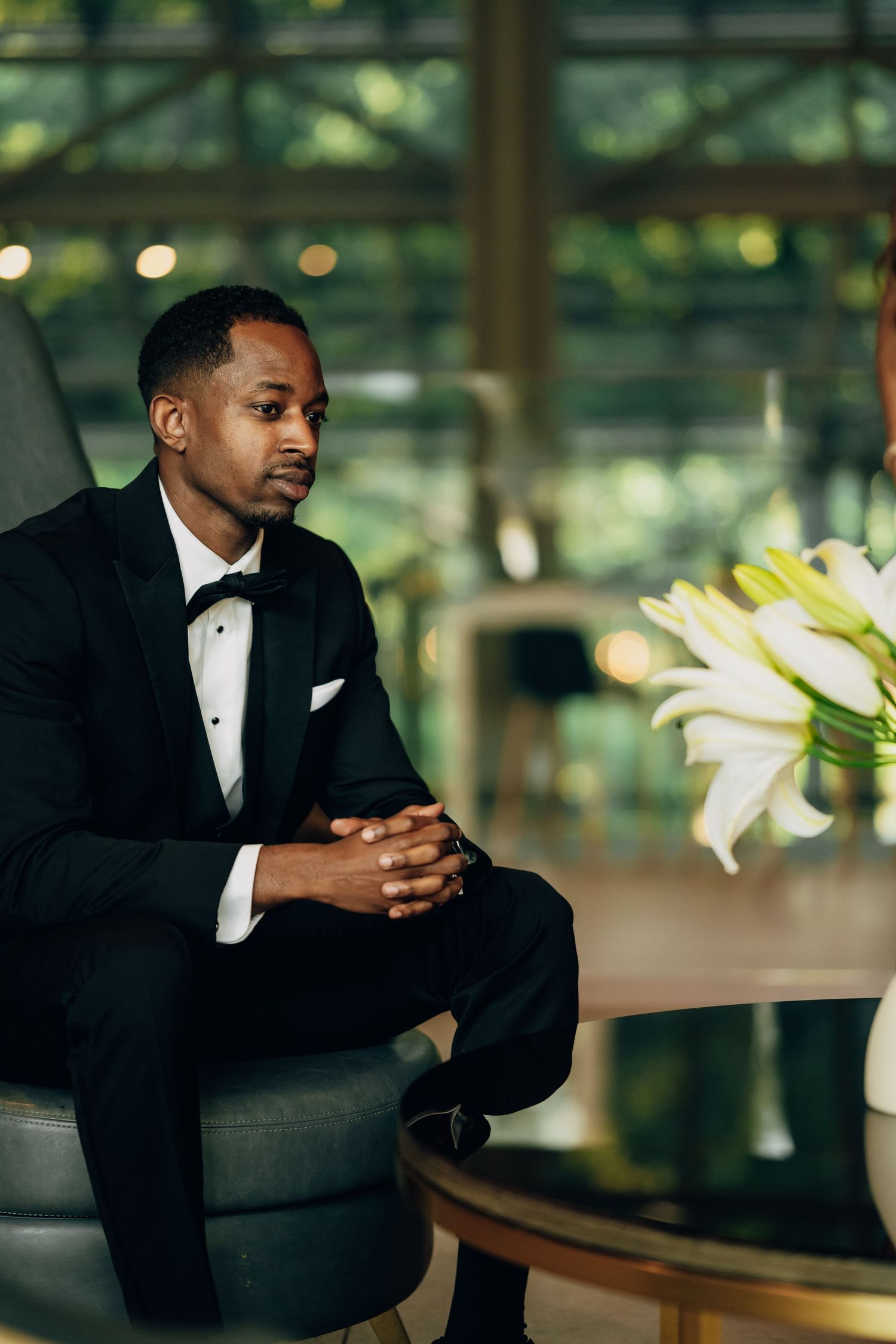 Man in tuxedo sitting thoughtfully in modern space with white lilies in background.