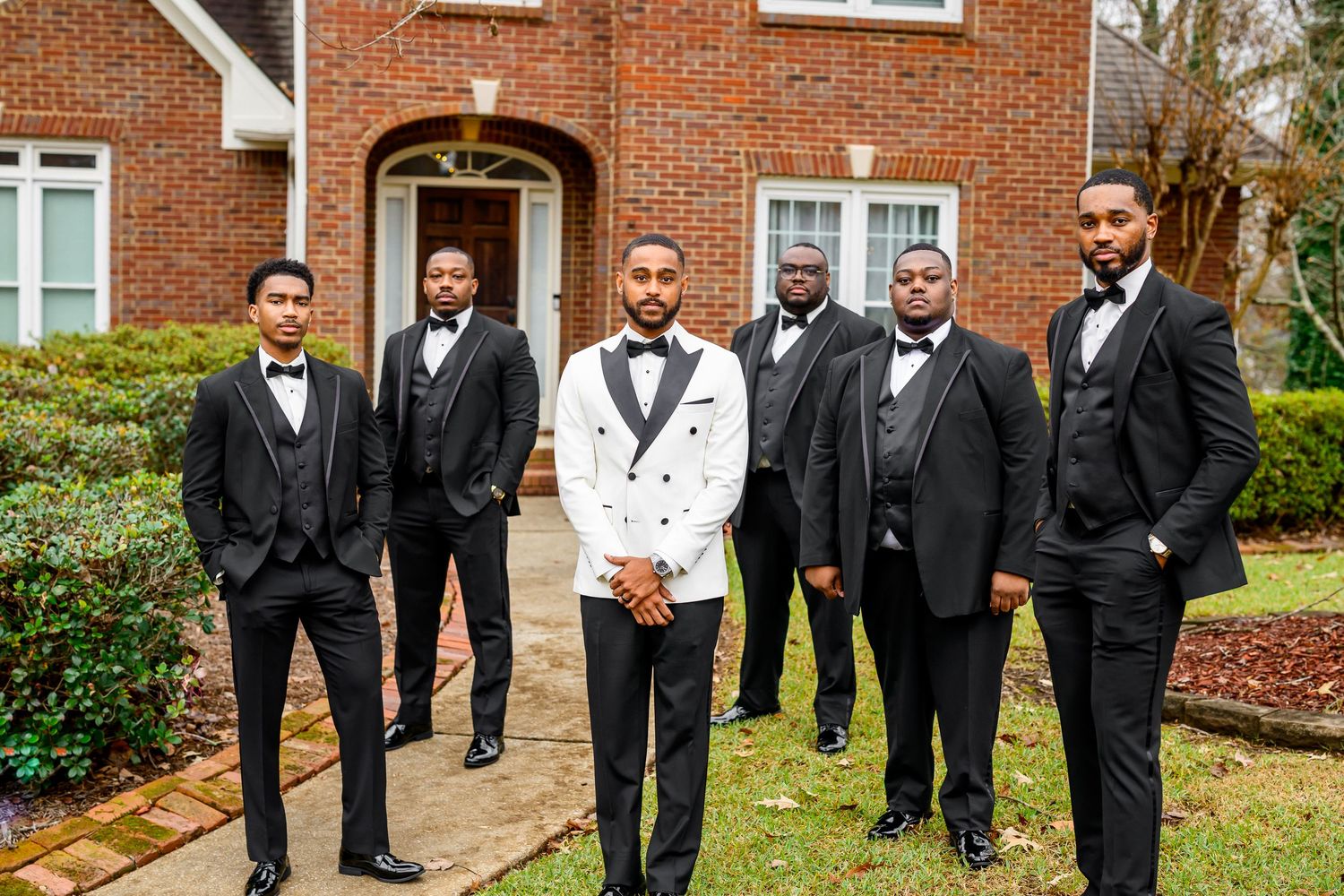 A group of groomsmen in black tuxedos pose together outside a brick building for wedding photos.