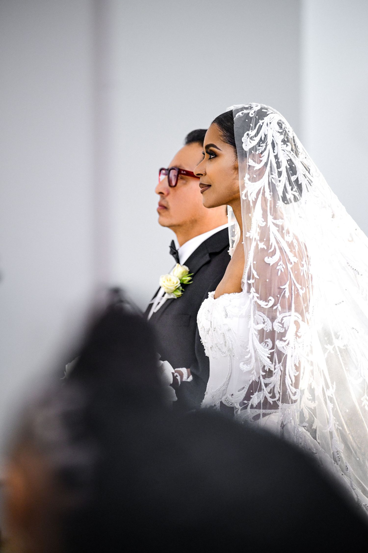 A bride and groom stand in profile during their wedding ceremony, with the bride wearing a delicate lace veil.