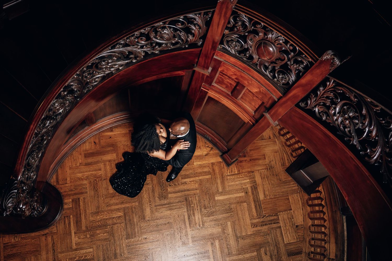 Overhead view of ornate wooden staircase with decorative railing in dark lighting.