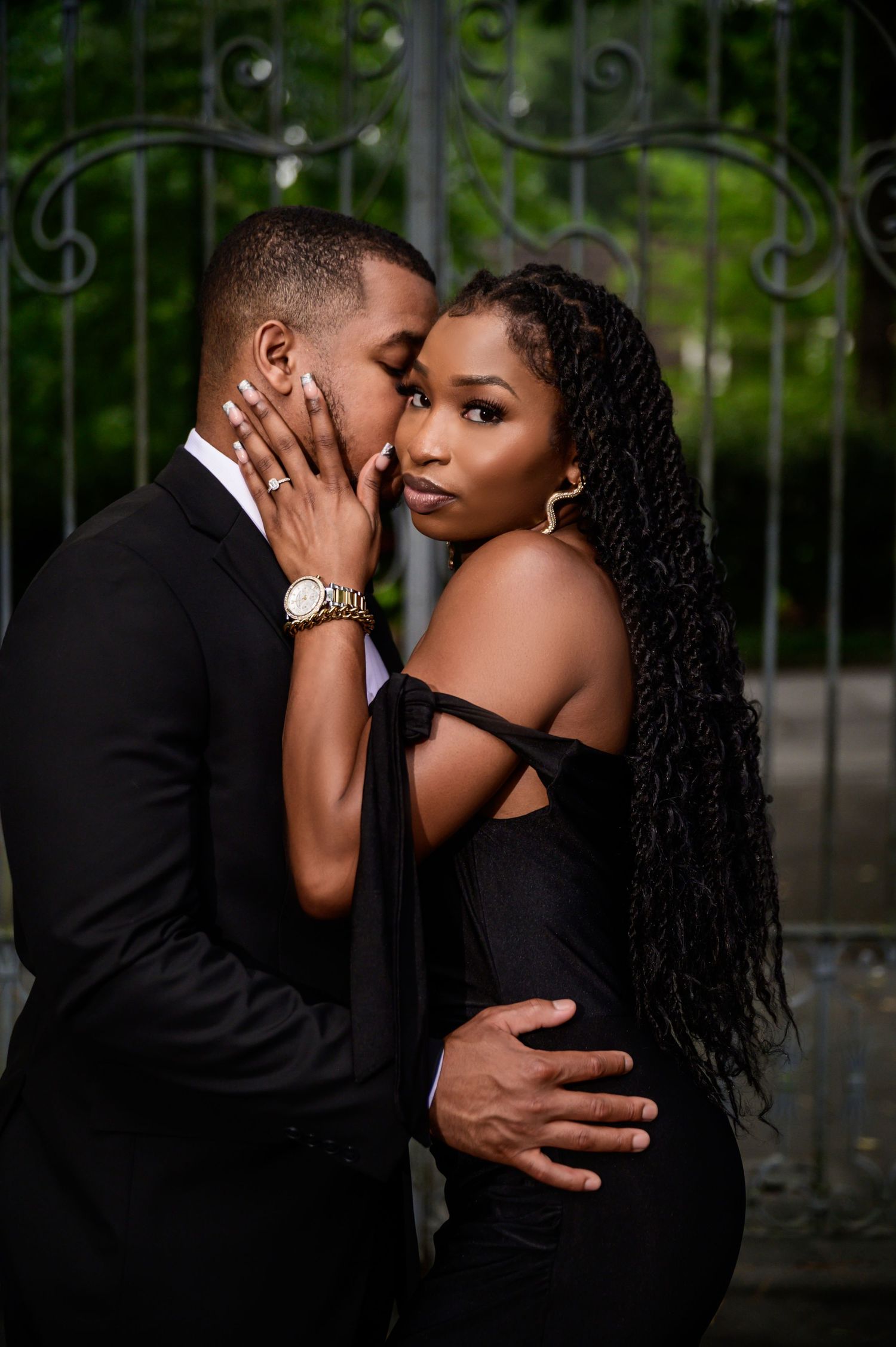 Intimate couple in black formal wear embrace tenderly against an outdoor backdrop.