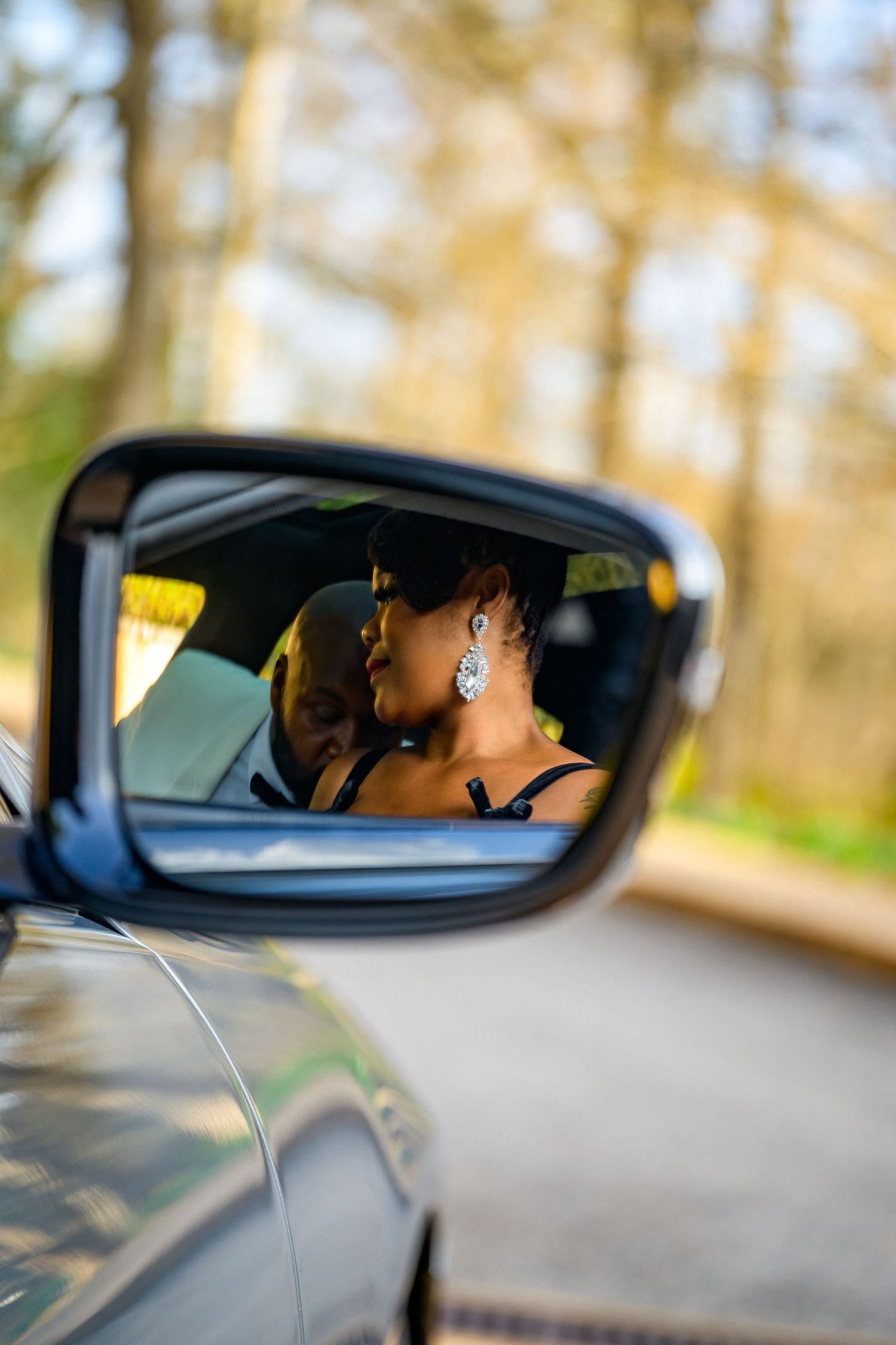 Car side mirror reflection shows driver in blurred bokeh effect on sunny autumn day with trees in background.