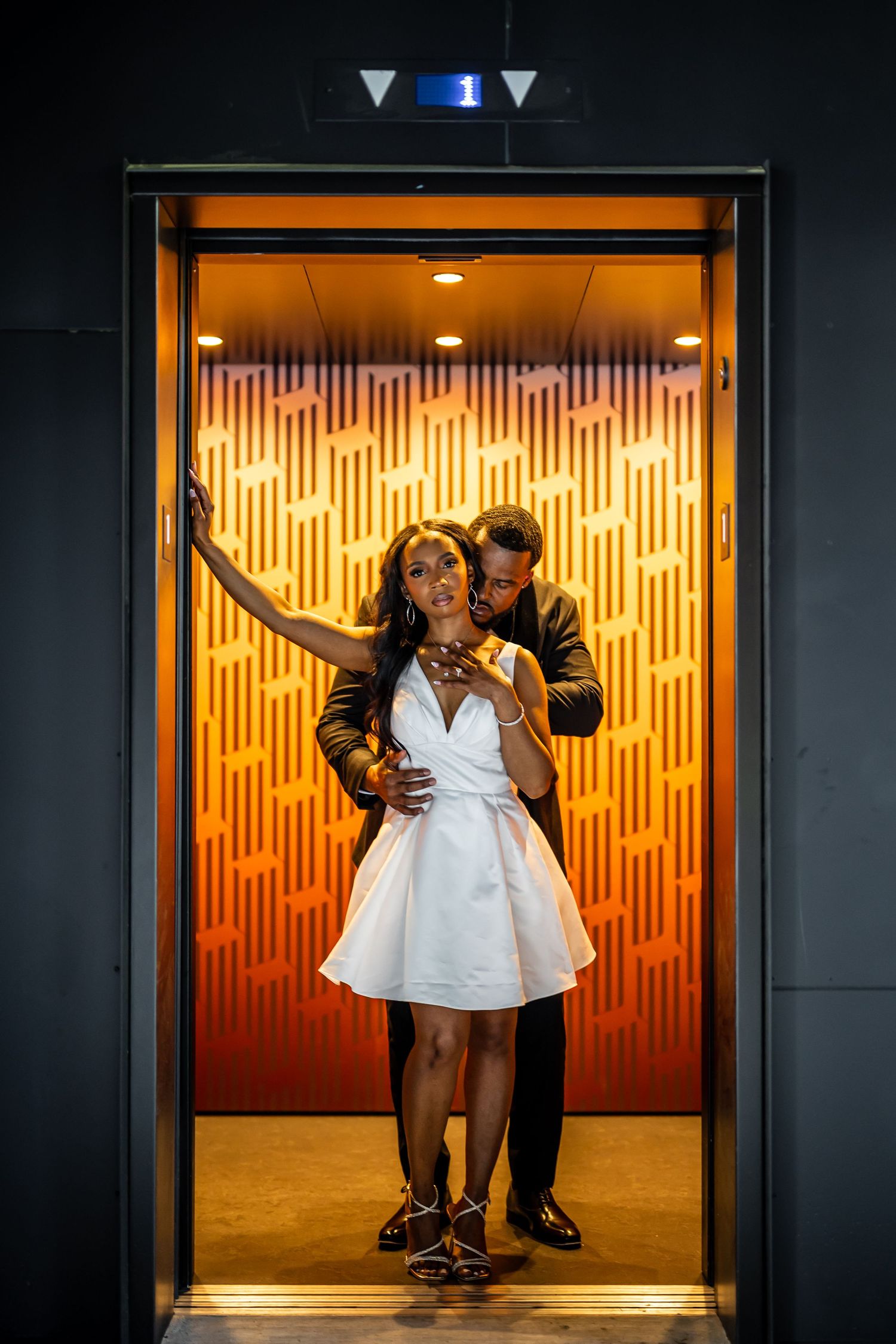 A romantic couple poses in an elevator with warm orange lighting and geometric wall patterns.