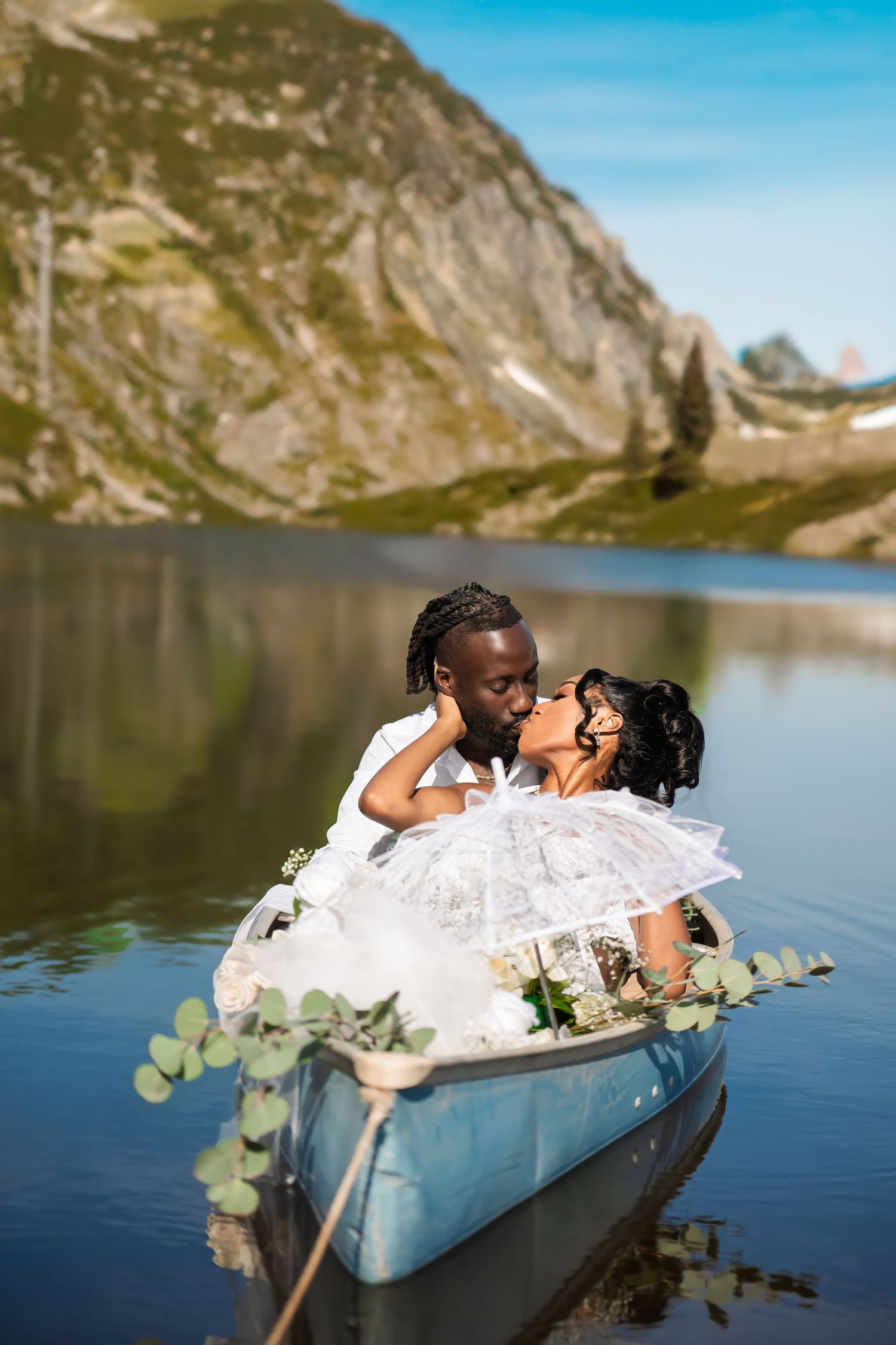 Newlyweds share romantic moment in blue canoe against backdrop of mountain lake.