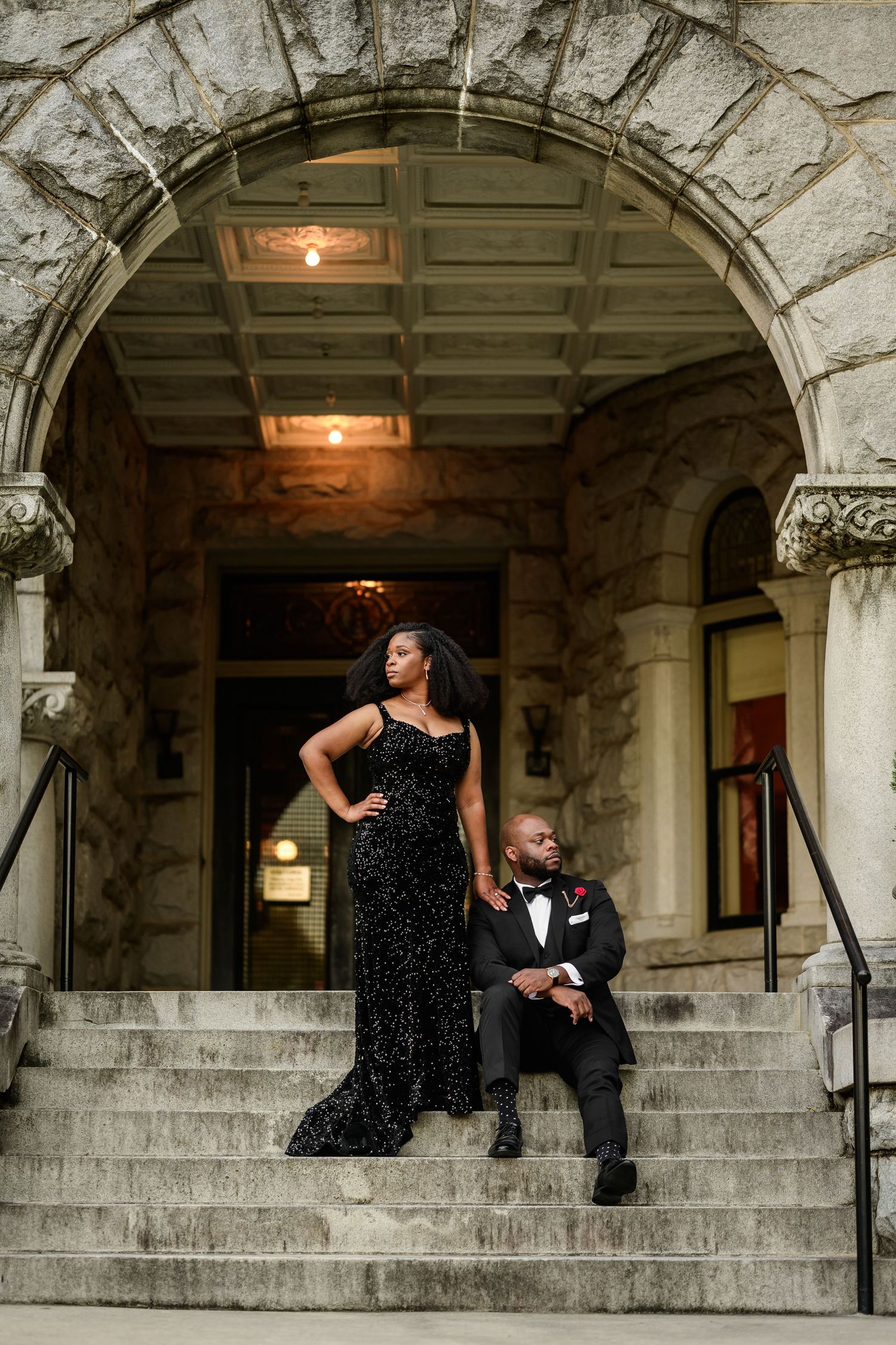Romantic photoshoot featuring couple in formal wear on grand stone steps with arched architecture.