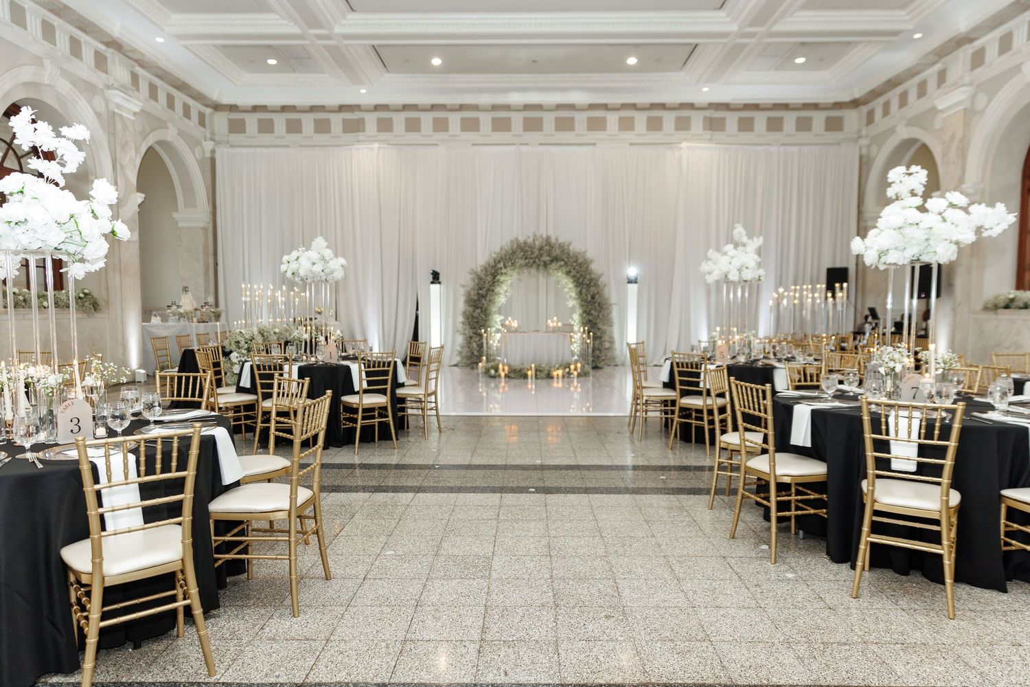 Luxurious wedding reception hall with white decor, chandeliers and gold chiavari chairs.