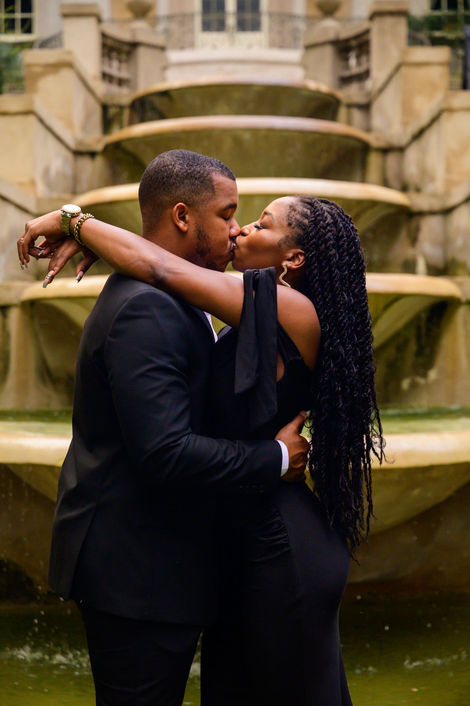 Romantic couple shares tender moments by ornate stone fountain in formal garden setting.