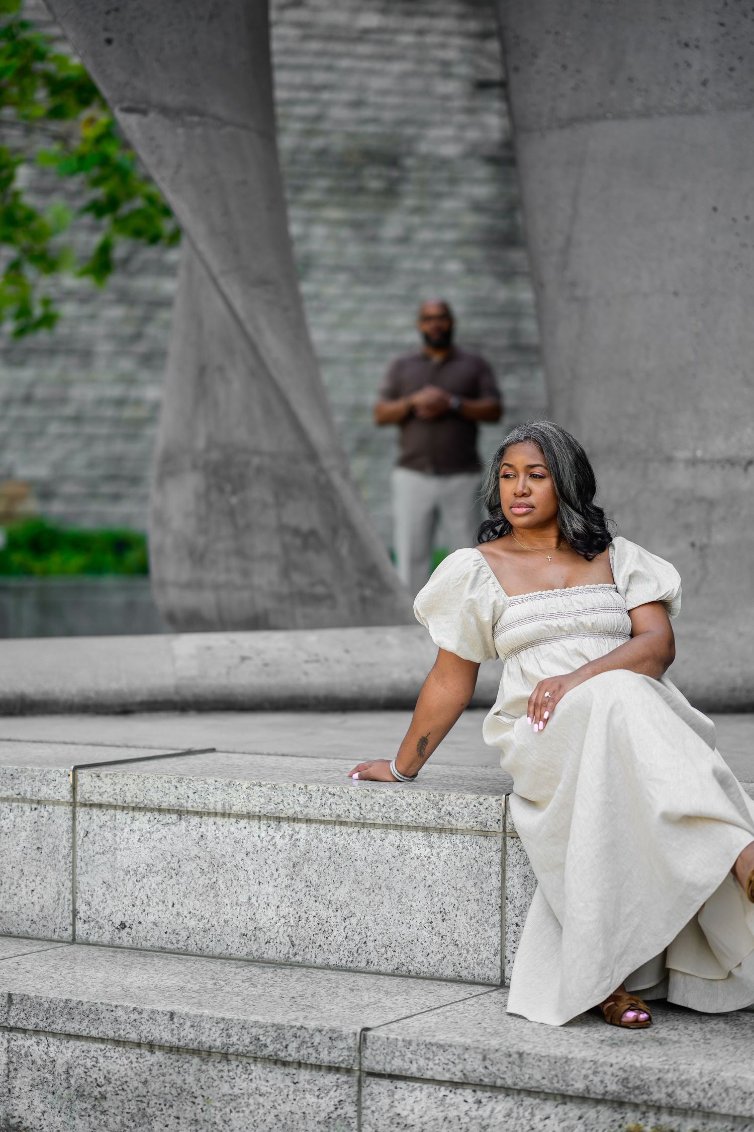 Person in white dress poses elegantly on concrete steps against stone wall backdrop.