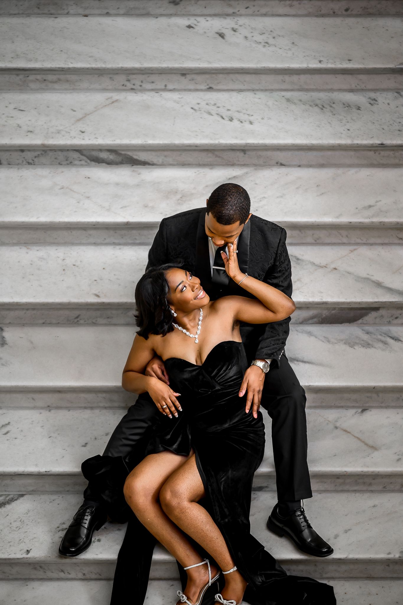 Elegant couple in black formal attire pose romantically on concrete steps during a photoshoot.