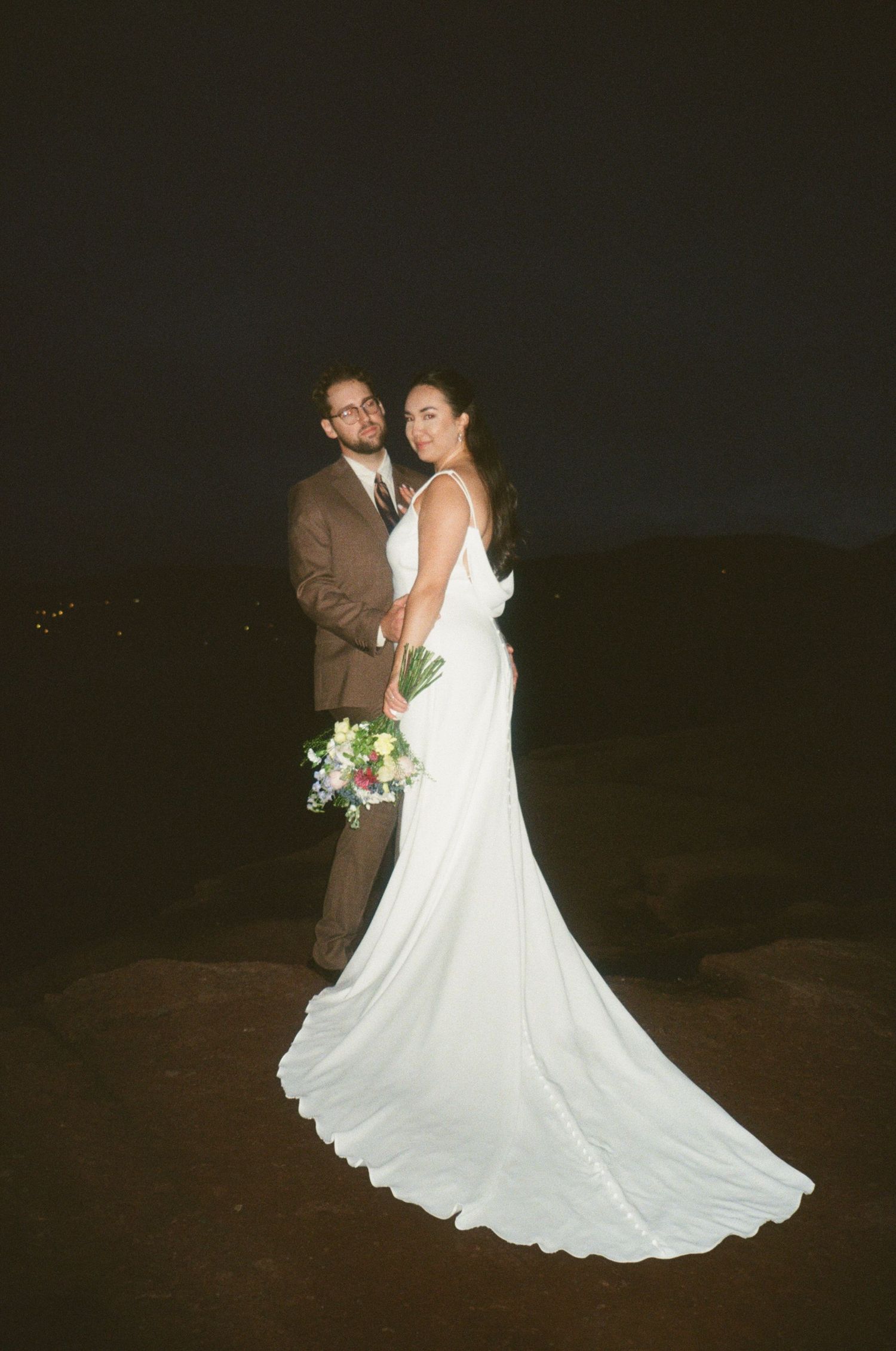 A bride in a flowing white gown and groom in brown suit pose together at night for a romantic wedding portrait.