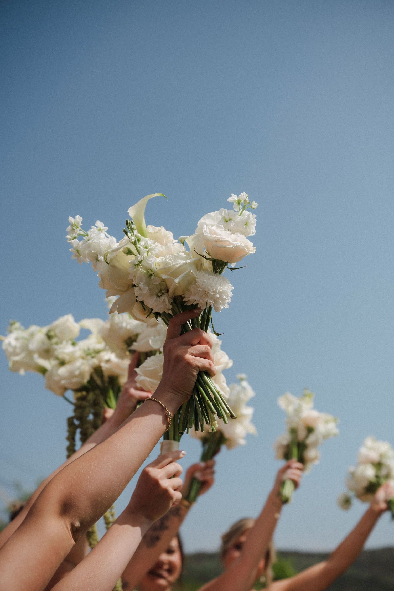 Bridesmaids holding white flower bouquets up against a clear blue sky at an outdoor wedding celebration.