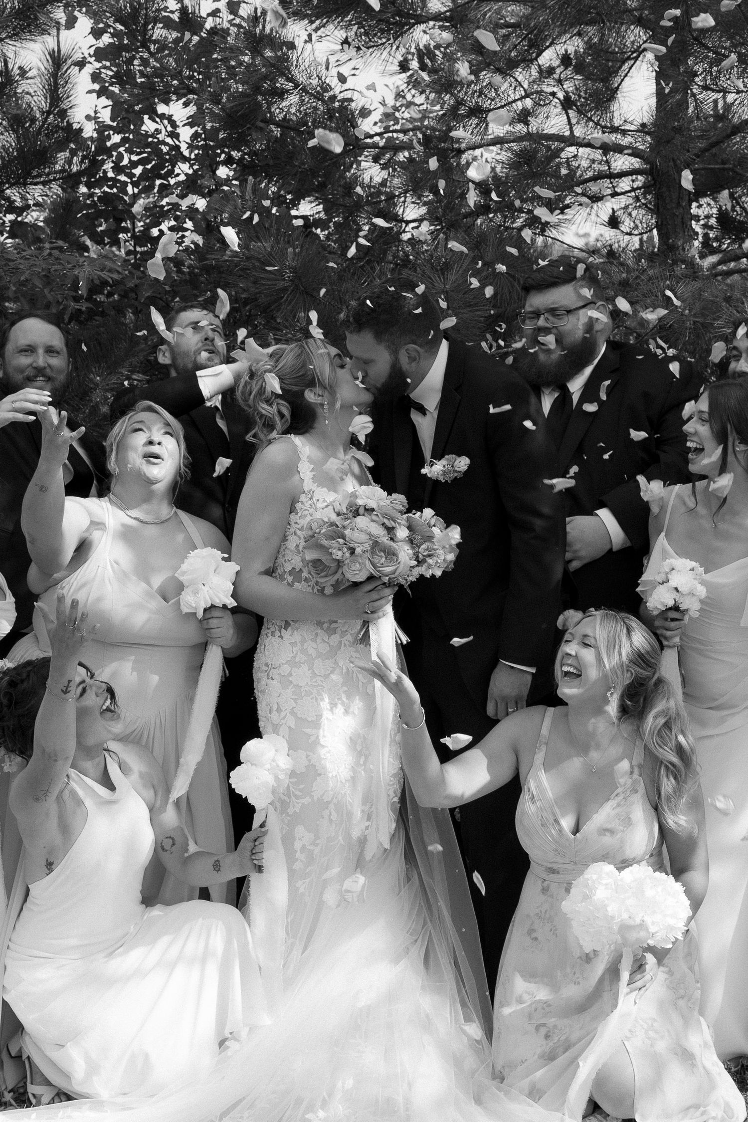 Black and white photo of bride and groom kissing surrounded by wedding party throwing confetti outdoors.