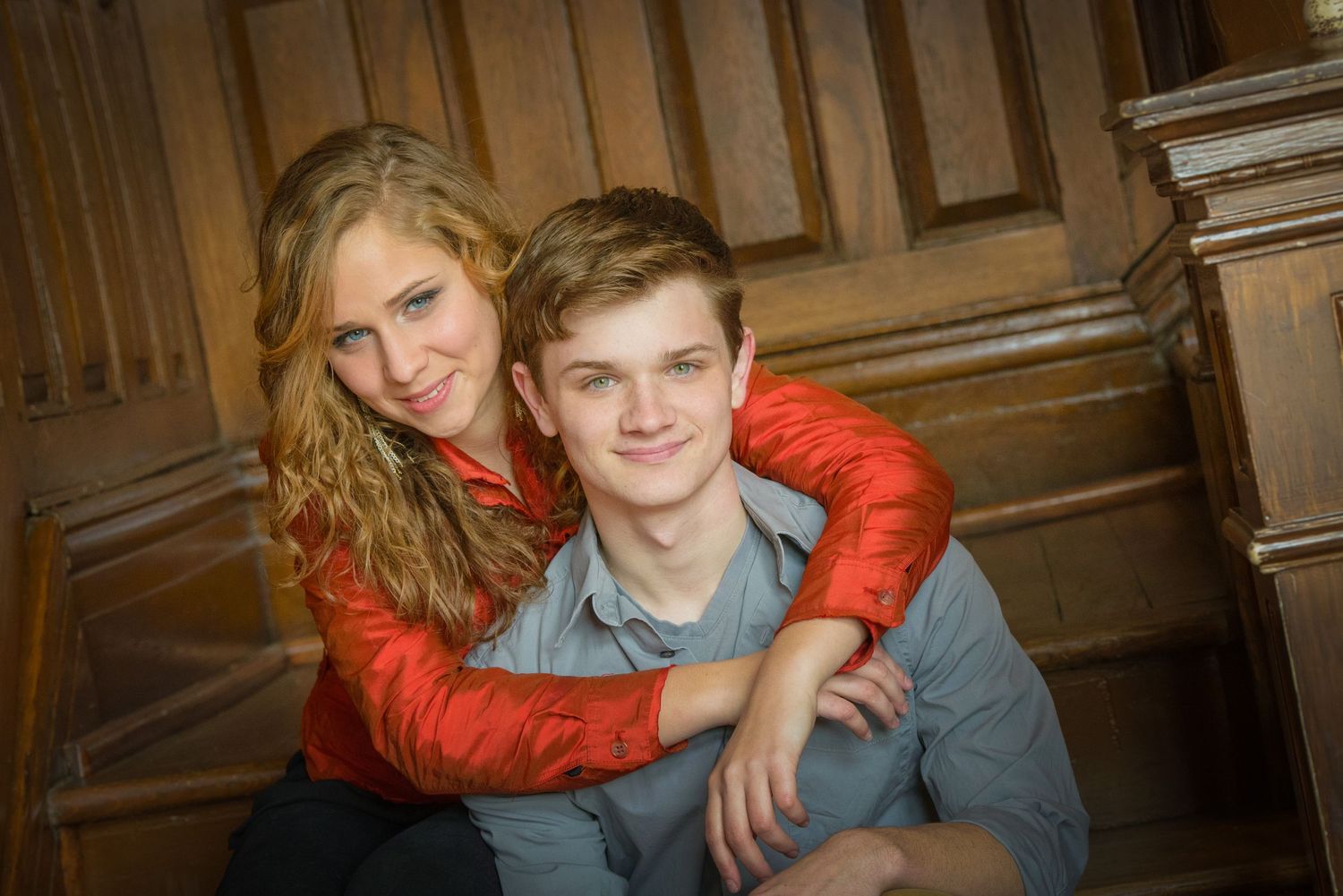Two people pose together in front of wood paneled wall, one wearing a bright orange jacket embracing the other from behind.