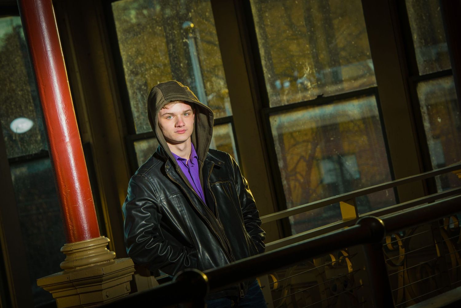 Figure in a black leather jacket and cap standing near a red pillar in a dimly lit industrial setting.