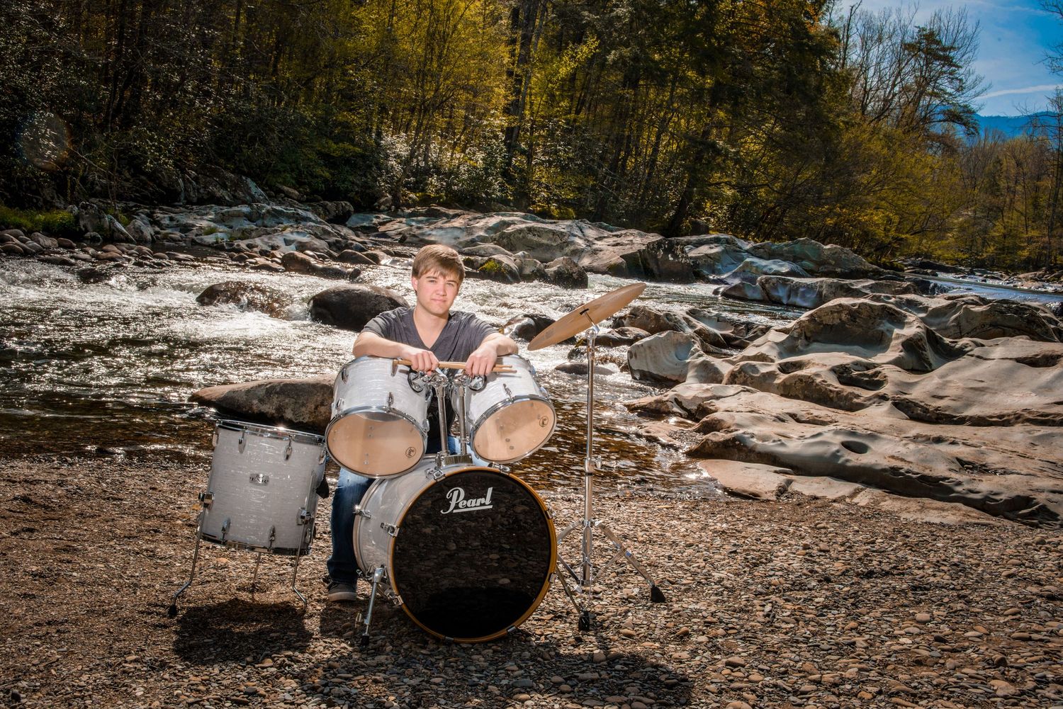 A drummer plays on a Pearl drum kit set up on rocky riverbank surrounded by autumn trees and flowing water.