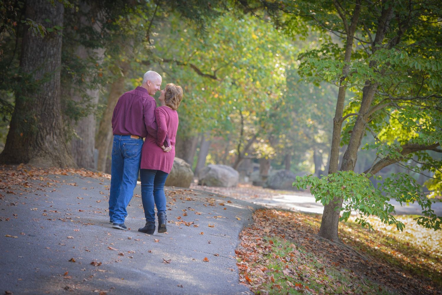 A couple in warm clothing stroll together on a tree-lined autumn path with fallen leaves scattered on the ground by Gatlinburg Photographers