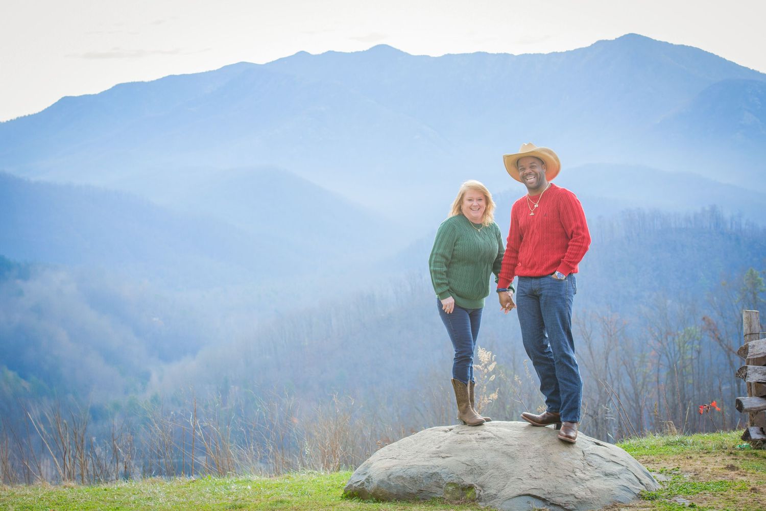 A couple stands together on a large rock overlooking misty blue mountains in the distance on a sunny day by Gatlinburg Photographers TN.