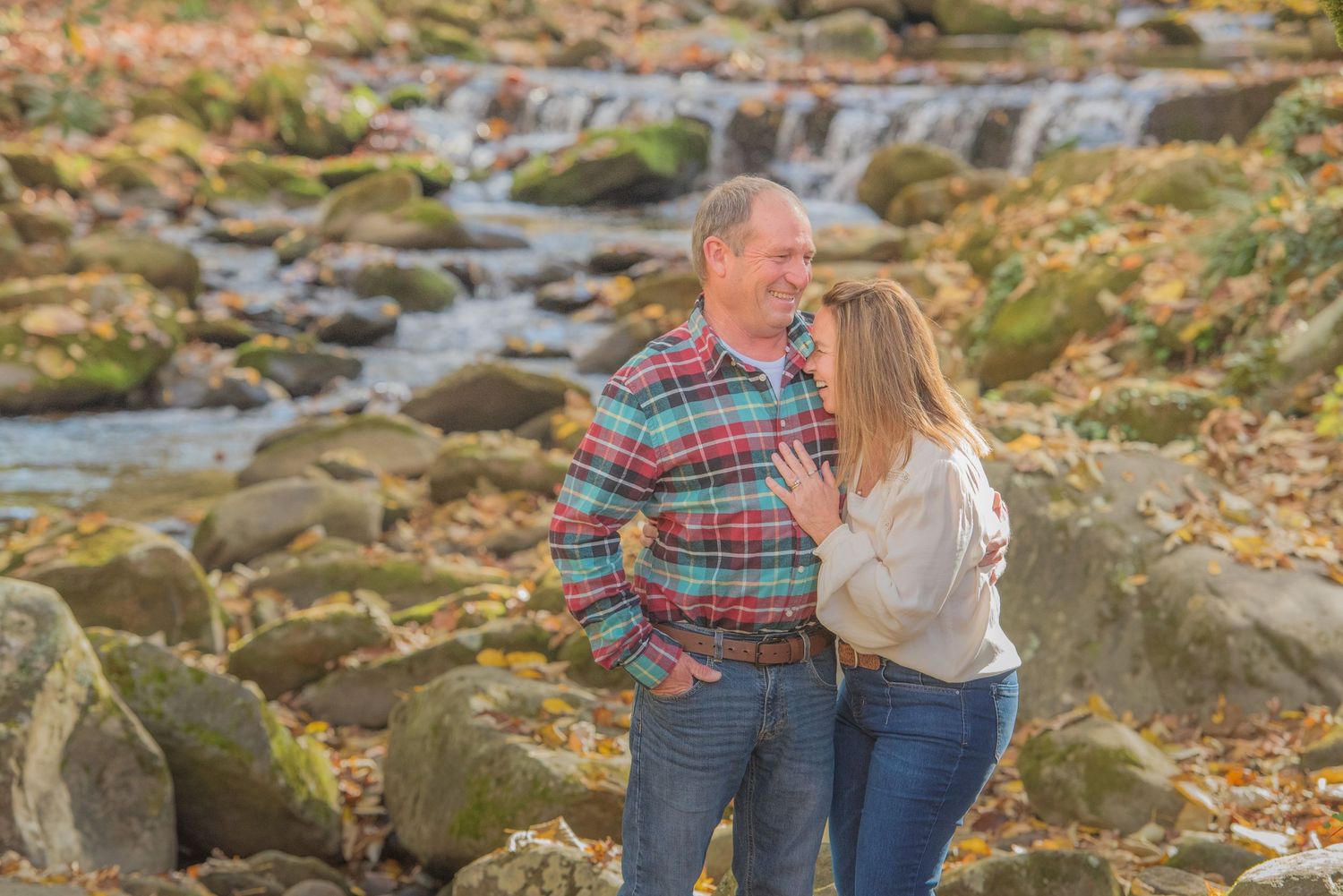 An anniversary couple embraces while standing together on mossy rocks near a cascading waterfall during autumn.