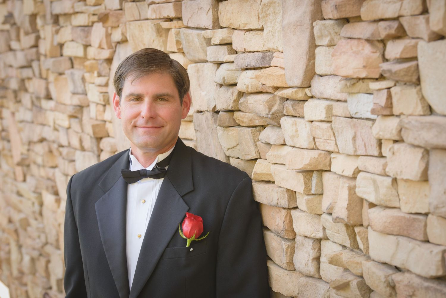 A groom in a black tuxedo with red boutonniere stands against a textured stone wall by Pigeon Forge Photographers.